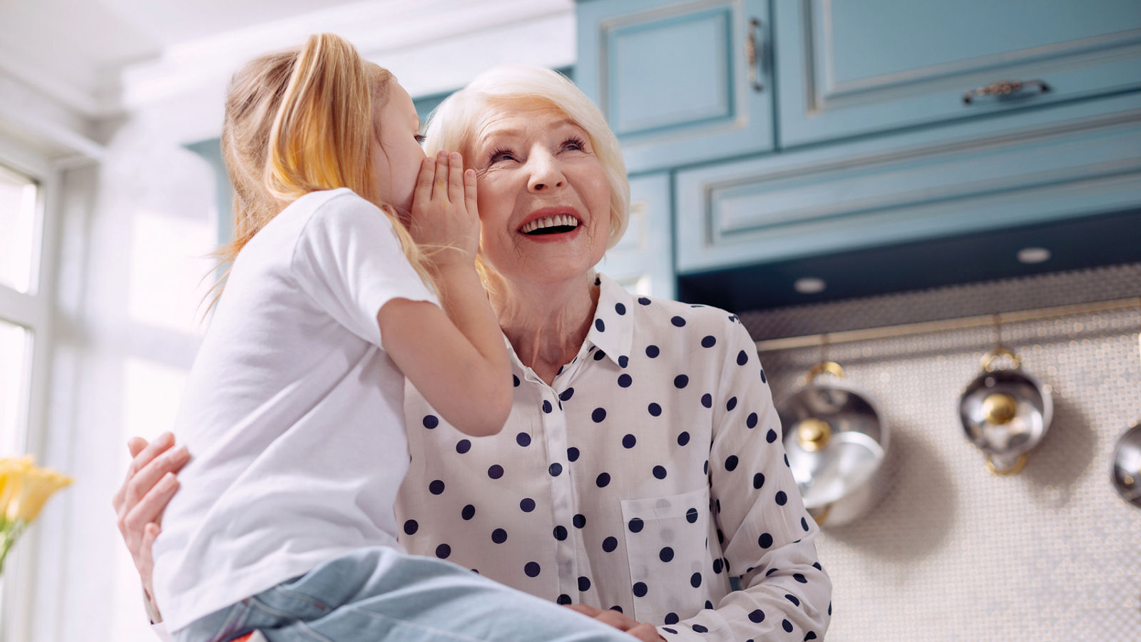 Our little secret. Pleasant charming elderly woman hugging her little granddaughter and listening to her secret while the girl sitting on the kitchen counter and whispering into her ear