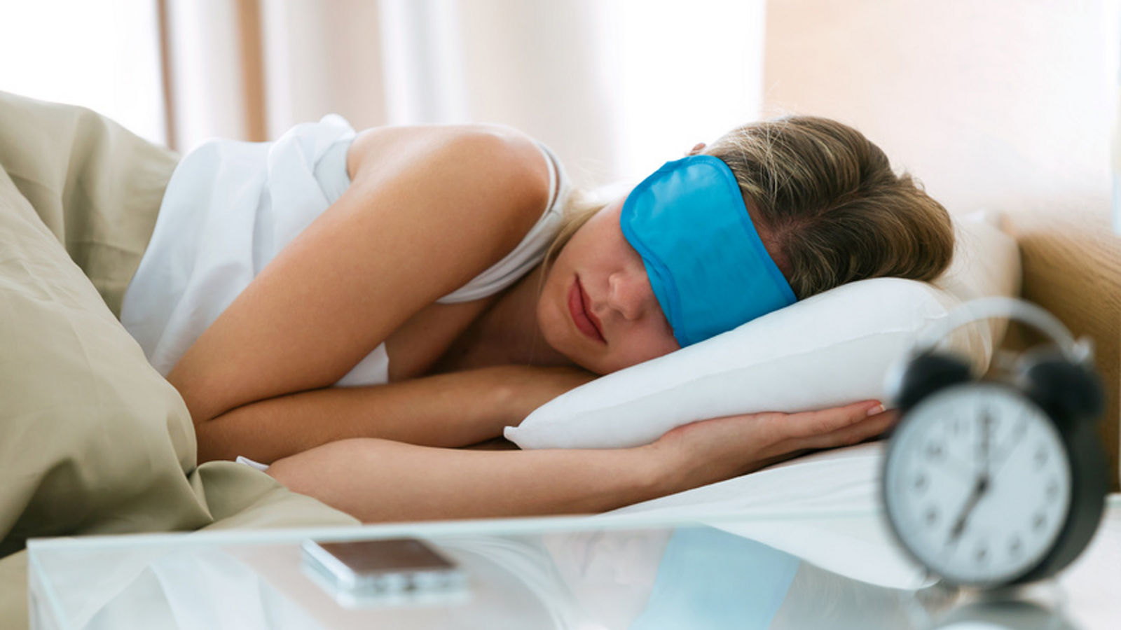 Beautiful young sleepy woman with sleep mask resting in bed with alarm clock in bedroom at home.