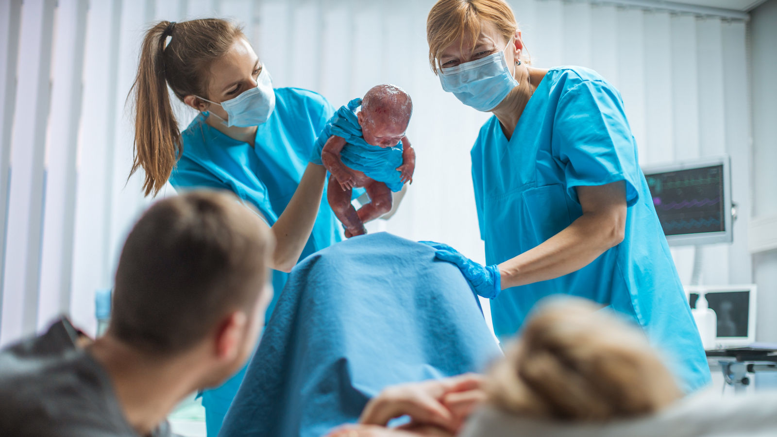 In the Hospital Midwive Holding Infant Baby. Woman in Labor Pushes and Gives Birth, Obstetricians Assist Delivery, Husband Holds Supports His Wife.