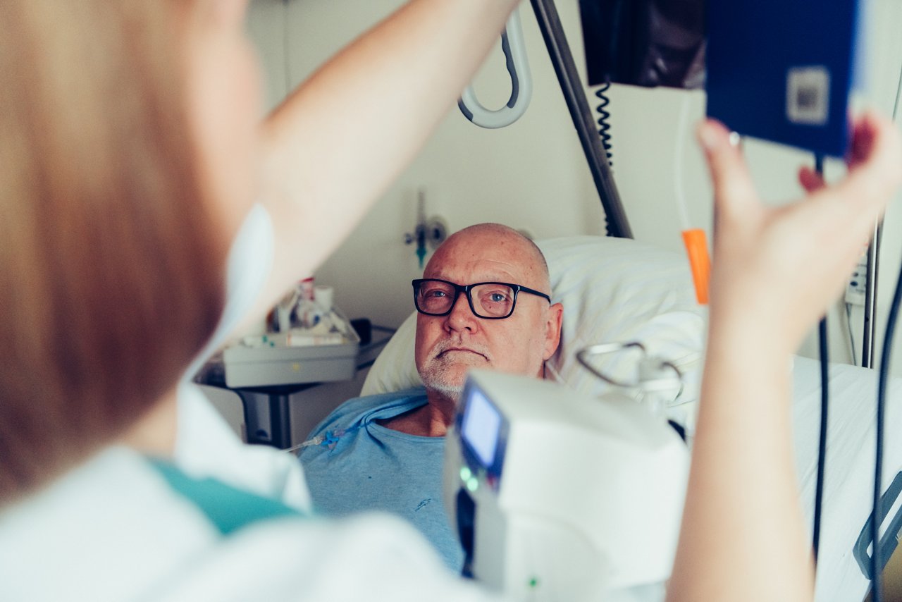 Patient liegt im Krankenbett und schaut der Pflegekraft dabei zu wie seine Chemotherapie vorbereitet wird