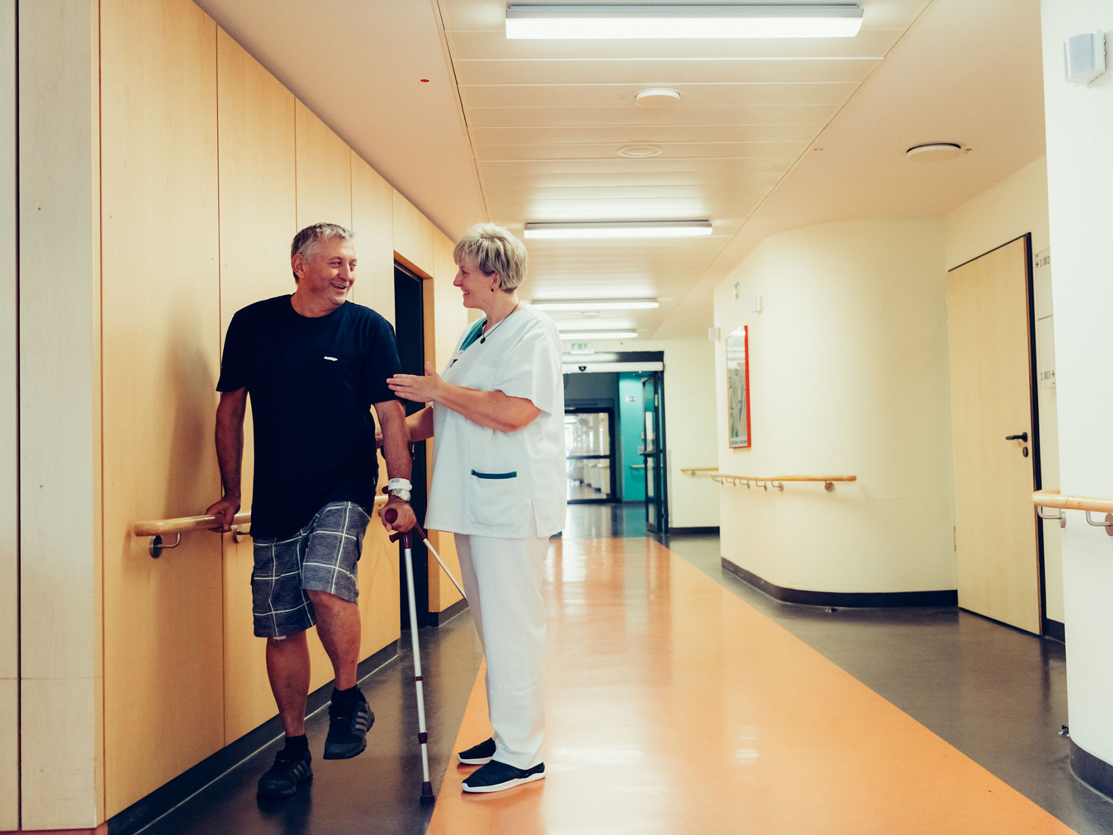 Patient mit Krücke hält sich am Handlauf fest und ist im Gespräch mit der Therapeutin