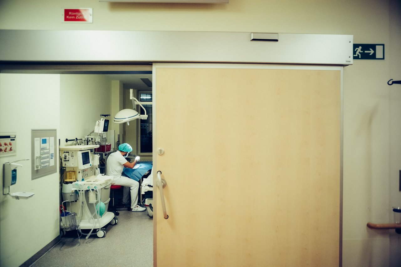 Blick durch eine offene Tür in einen Raum in dem eine Person mit OP-Haube und Mundschutz das Behandlungsbesteck prüft