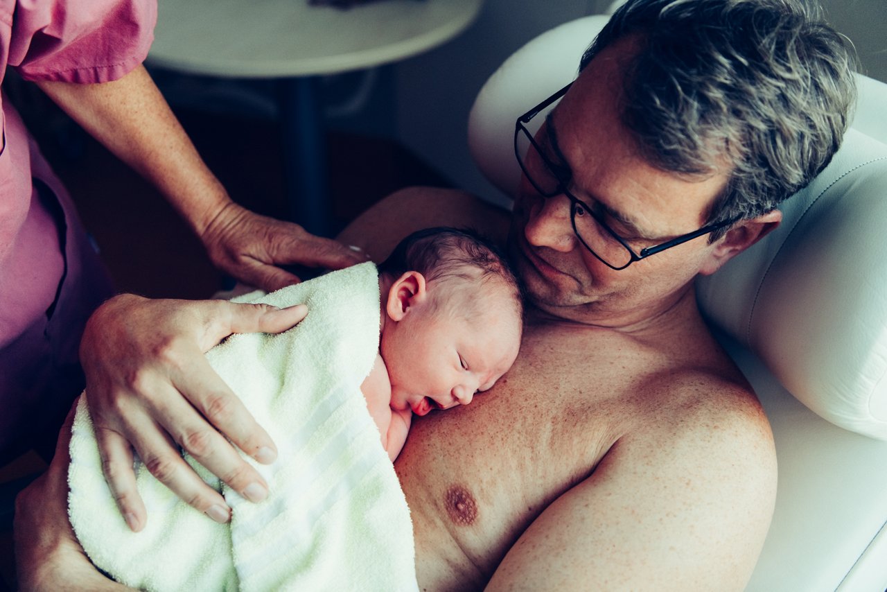 Vater hat Baby auf dem nacktem Oberkörper, im Anschnitt sieht man eine Hebamme