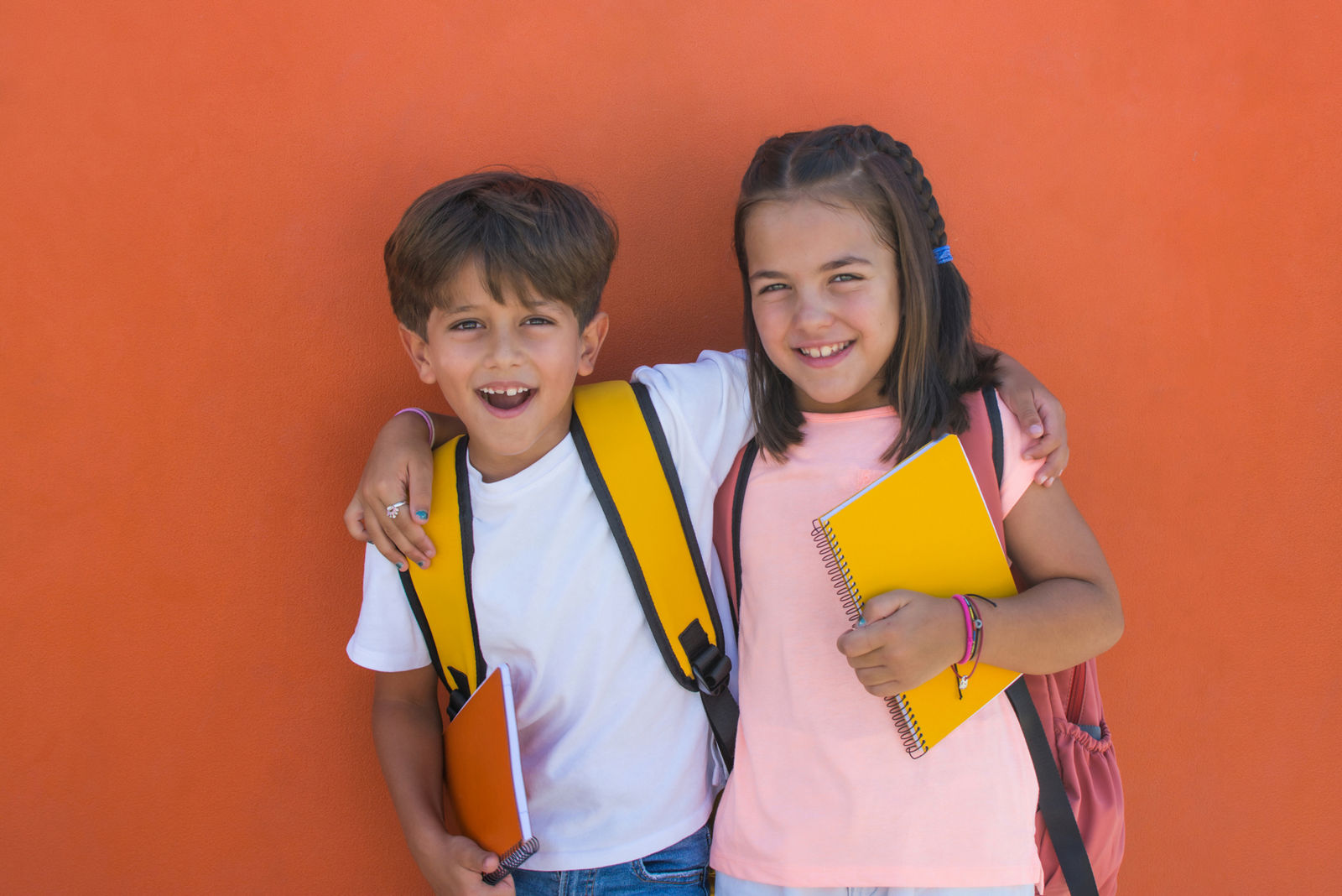 Elementary age children with facial expression of joy on the first day of school. Smiling preschool girl and boy of 7s with backpacks and notebook with orange background. Happiness concept