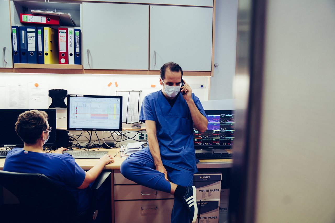 Pflegekraft lehnt an Schreibtisch und telefoniert, eine weitere Pflegekraft links davon arbeitet am PC