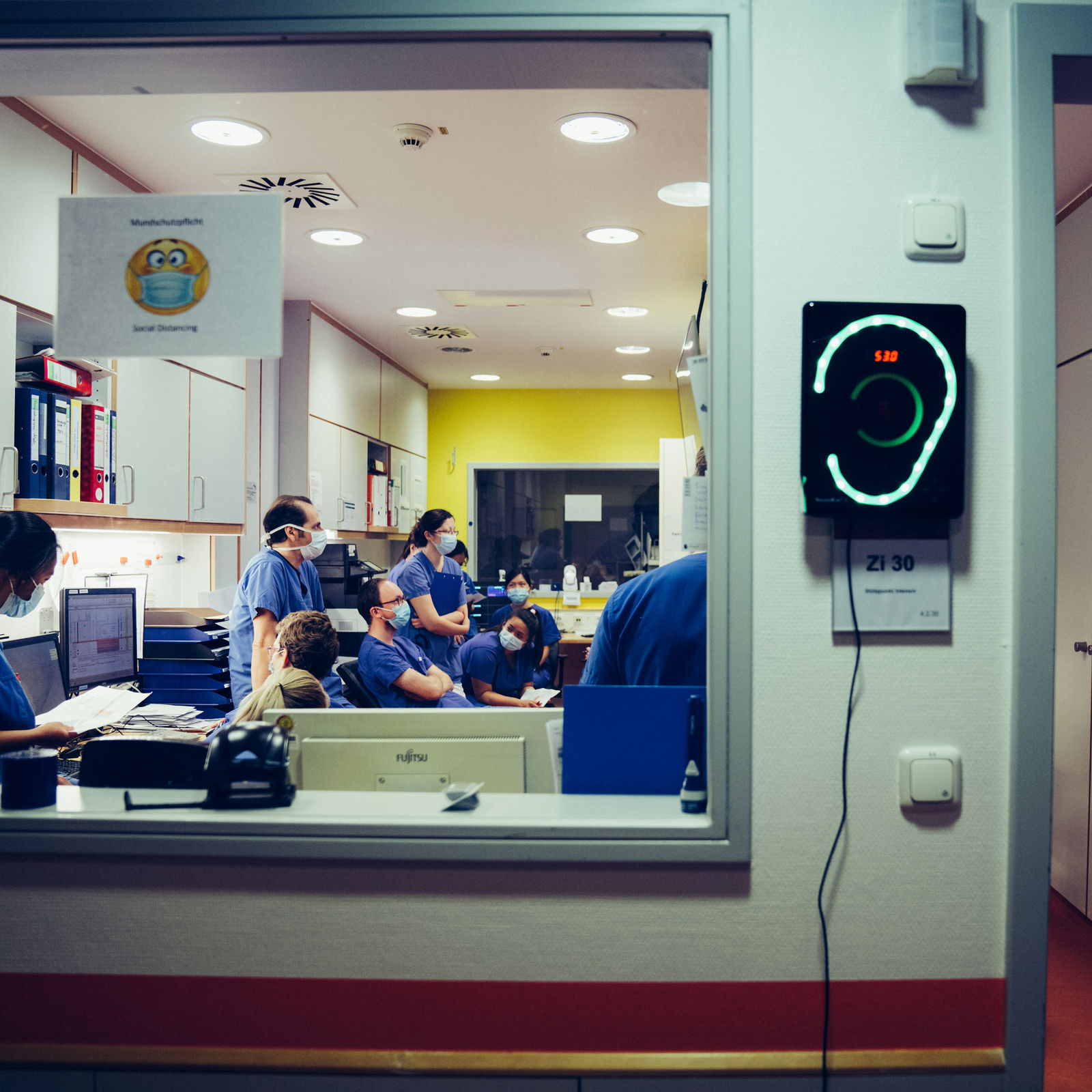 Blick ins Stationszimmer mit mehreren Pflegekräften in blauer Bekleidung und Mundschutz in einer Besprechung