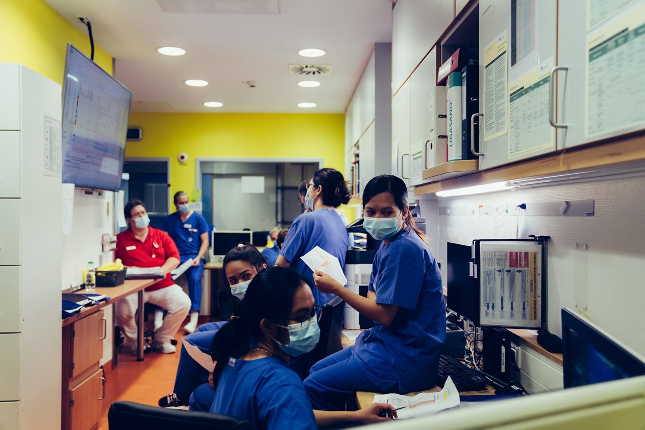 Mehre Pflegekräfte in einem Stationszimmer,  manche schauen auf einen Monitor, der links an der Wand hängt, Personen im Vordergrund scheinen auf einen PC-Bildschirm auf der rechten Seite zu blicken