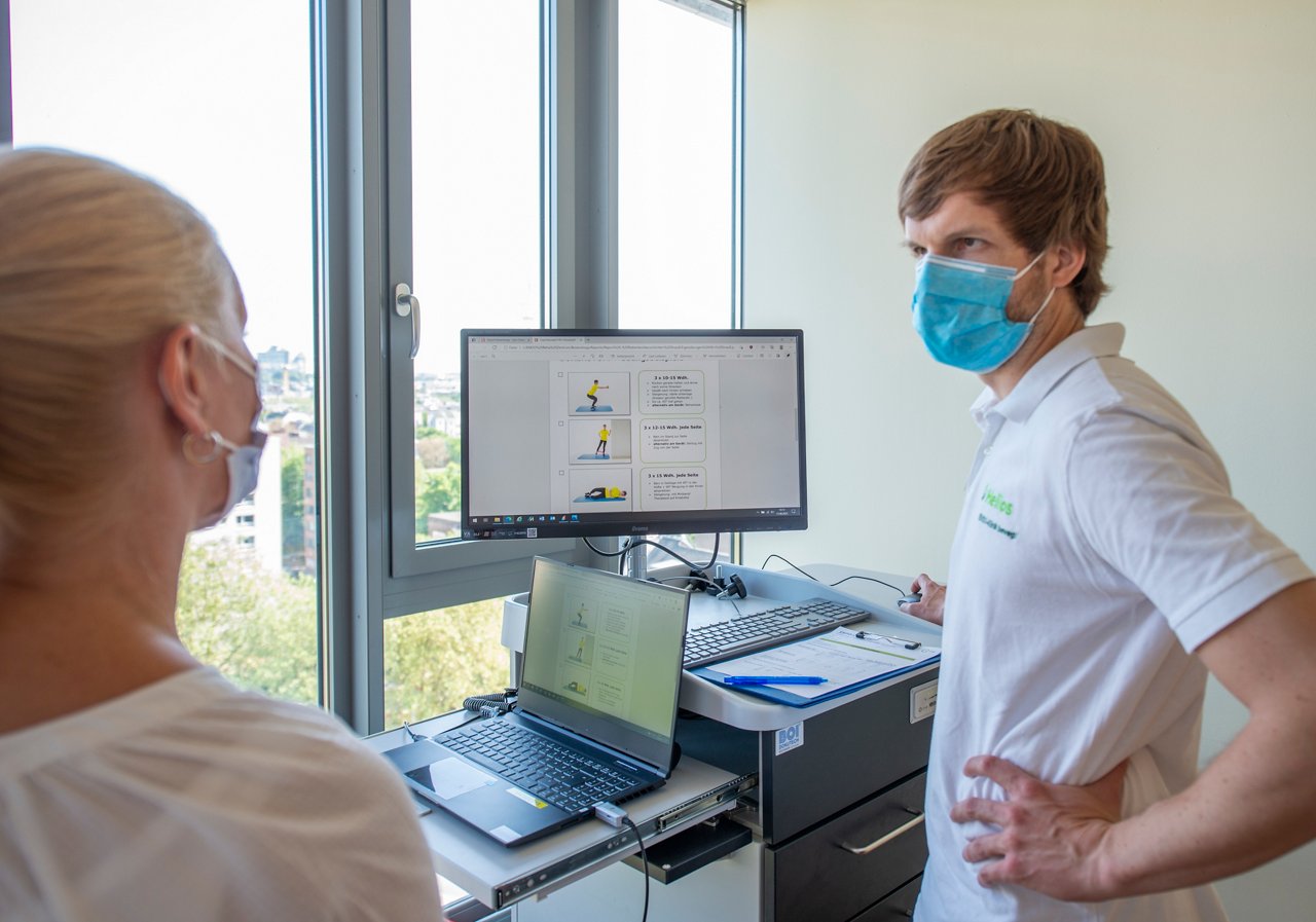 Physiotherapeut im GEspräch mit Patienten. Erklärung Therapie, mit Maske