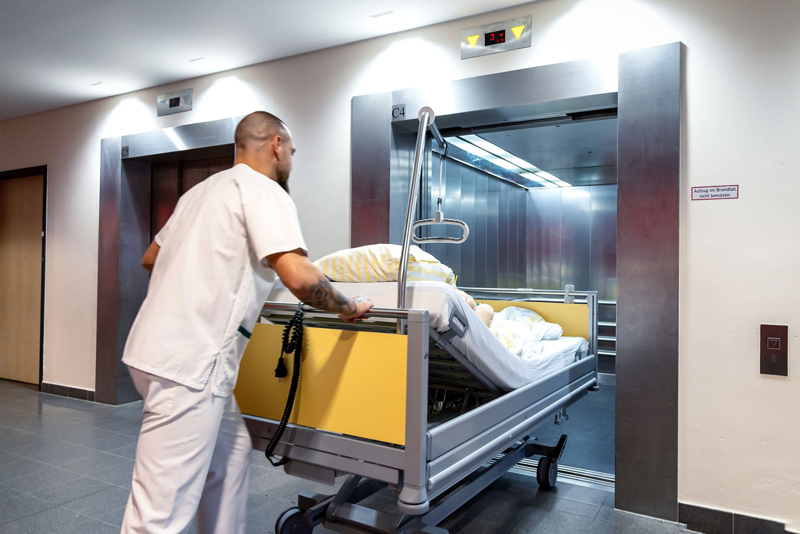 Krankenpflegehelfer Dominik mit Patientenbett am Fahrstuhl