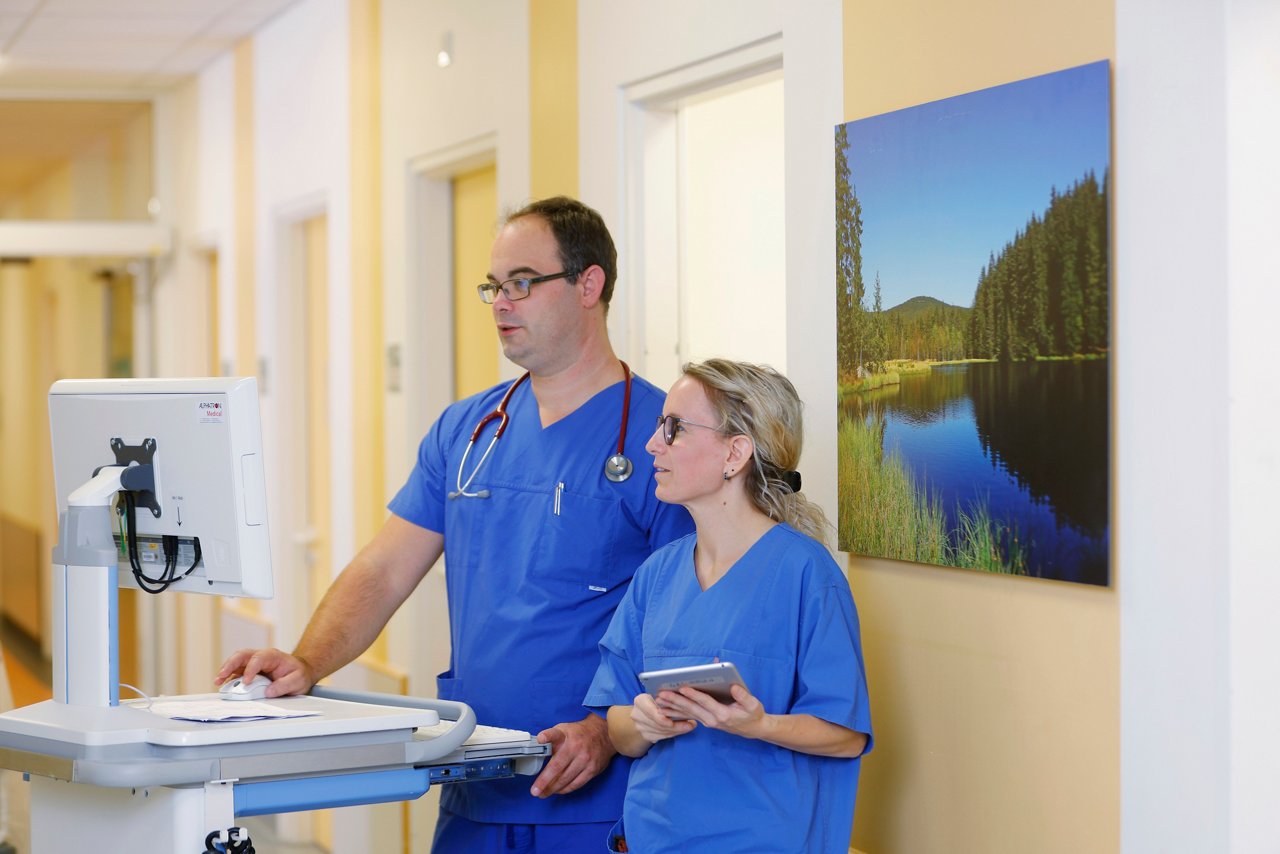 Frau und Mann in blauem Kasack im Gespräch vor Monitor