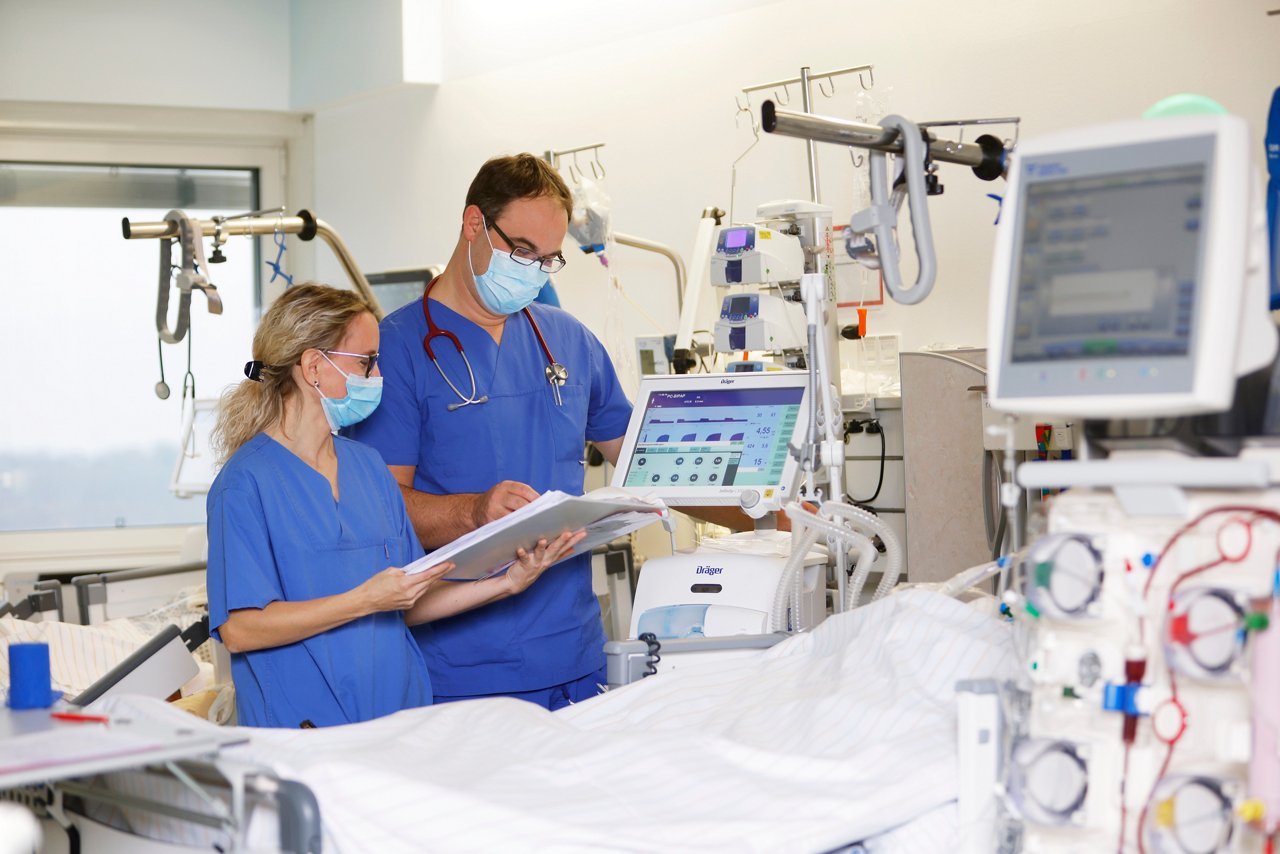 Zwei Personen in blauem Kasack blicken in einen Ordner neben leerem Patientenbett