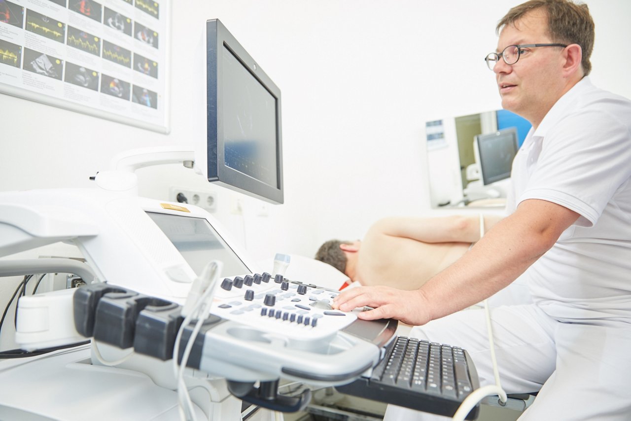  Arzt vor Ultraschallgerät mit Patient in Seitenlage im Hintergrund