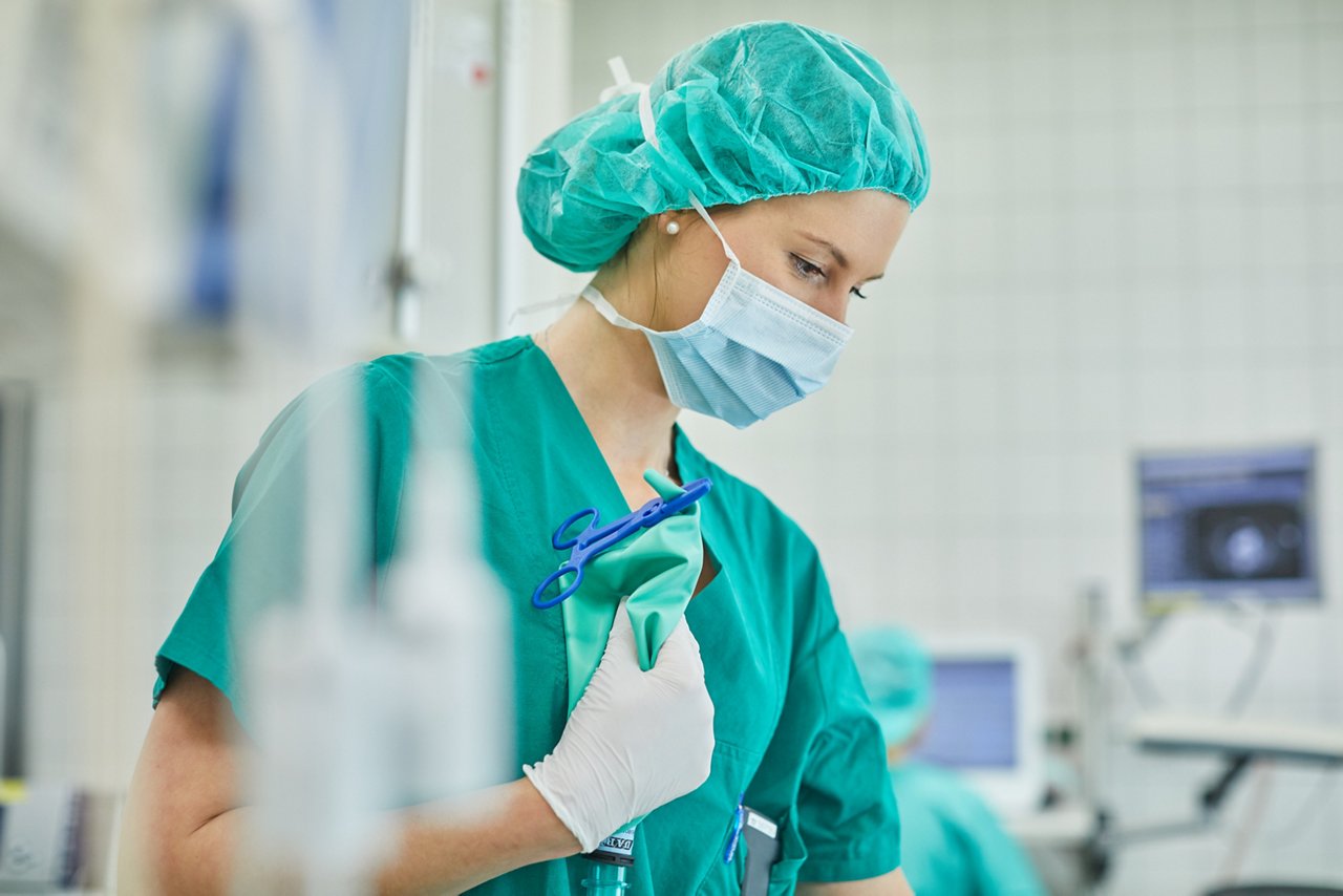 Frau in OP-Kleidung steht nach unten schauend im Operationssaal und hält einen Beutel mit Abklemmschere.