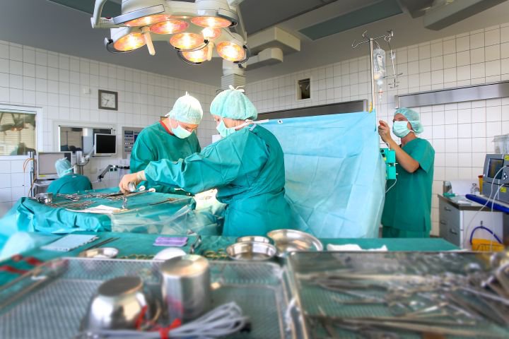 Operationssaal mit drei Menschen in ärztlicher Schutzausrüstung; im Vordergrund Operationsbesteck