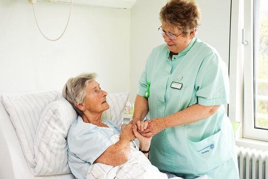 Grüne Dame hält aufmunternd die Hände einer Patientin im Patientenbett