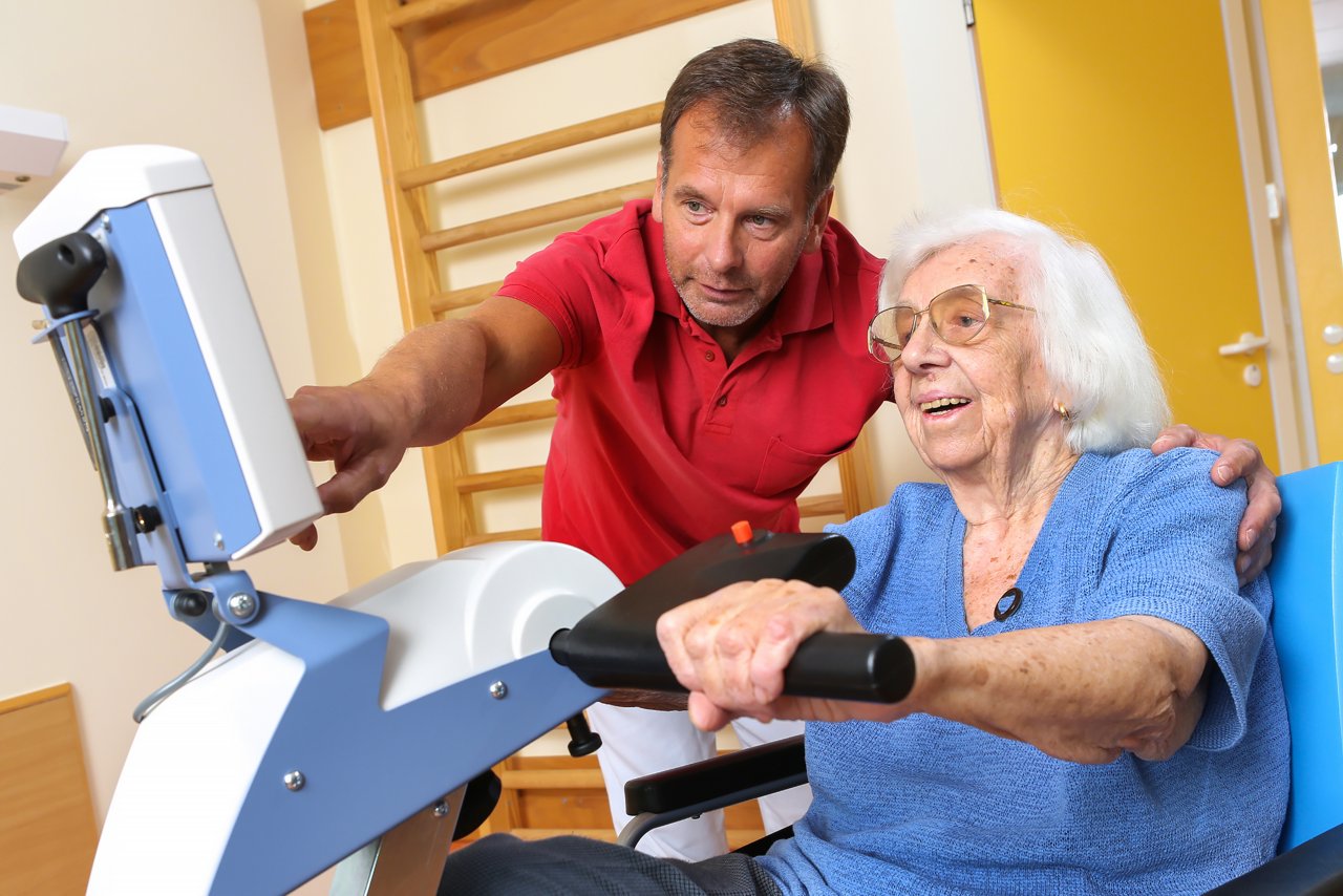Physiotherapeut zeigt älterer Patientin im Rollstuhl einen Monitor eines Behandlungsgerätes zum Treten
