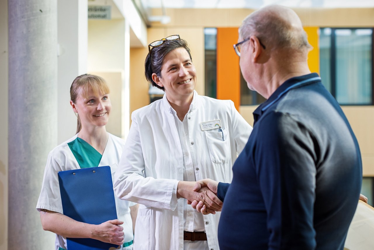 Patient und Ärztin schütteln die Hände. Pflegekraft steht mit Klemmbrett im Hintergrund