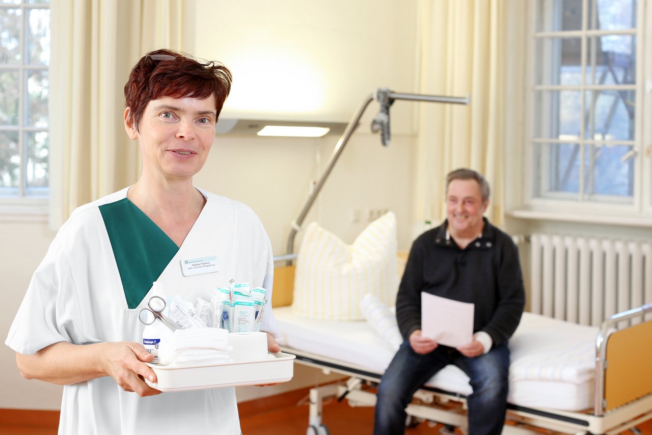 Die Pflegekraft steht mit Verbandsmaterial im Patientenzimmer der Station. Patient sitzt im Hintergrund auf dem Bett und hält einen Zettel in der Hand