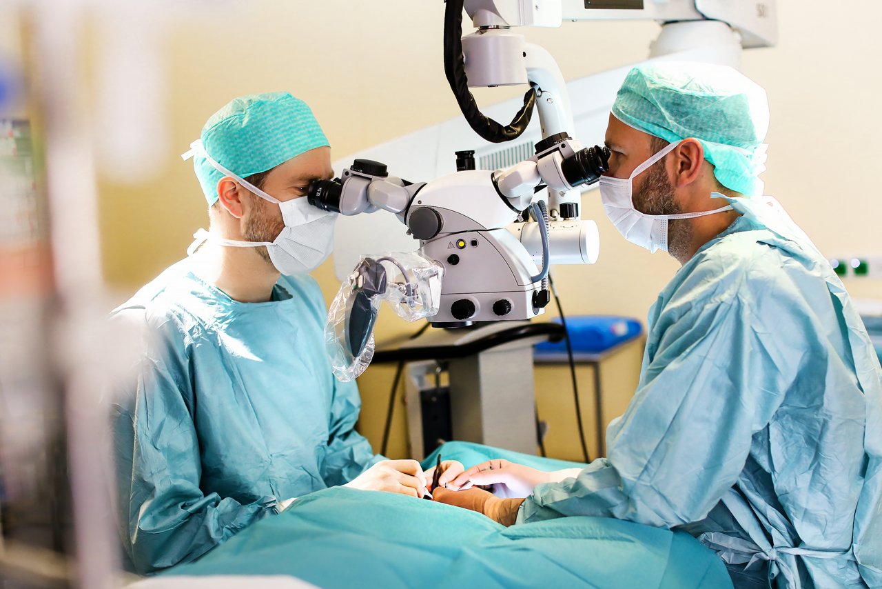 Zwei Operateure stehen im Operationssaal und schauen während der Operation durch ein Mikroskop. Sie halten Operationsbesteck in den Händen. 