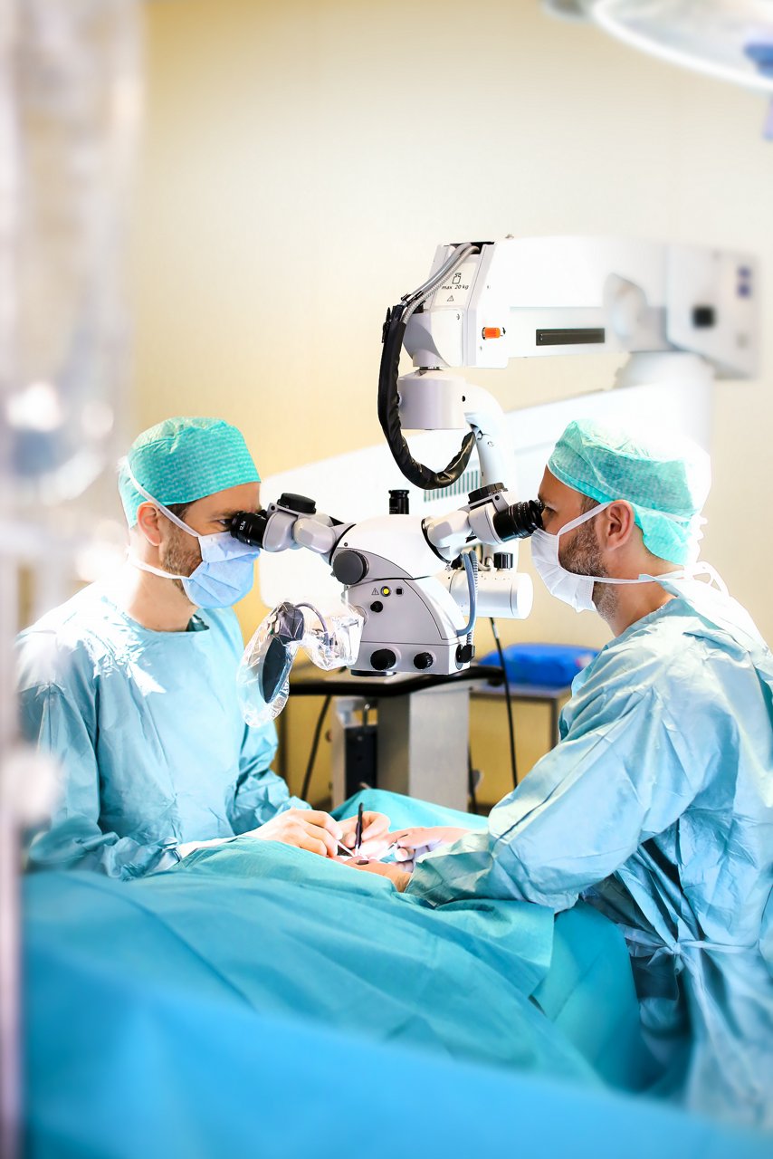 Zwei Operateure stehen im Operationssaal und schauen während der Operation durch ein Mikroskop. Sie halten Operationsbesteck in den Händen. 