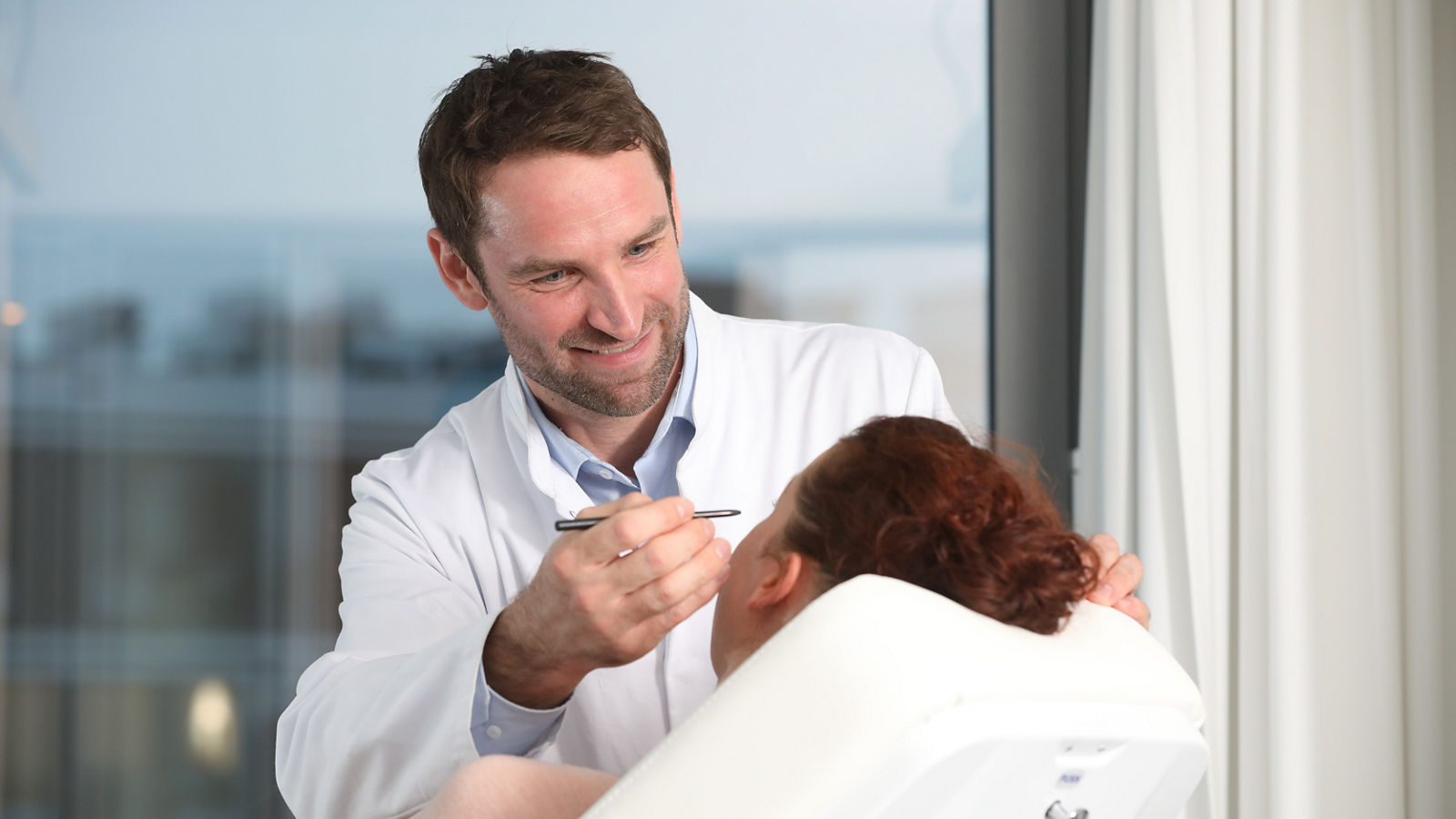 Arzt des plastisch-chirurgischen Fachbereiches zeichnet Korrekturlinien im Gesicht einer Patienten, die auf einem Behandlungsstuhl liegt.. 