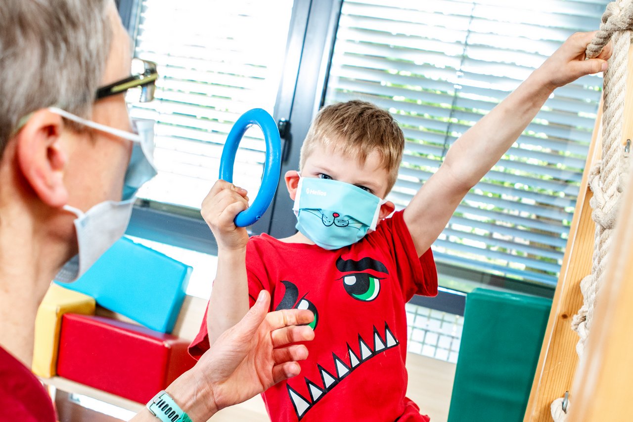 Kleiner Junge mit Katzenmaske bei der Bewegungstherapie mit Ärztin. Der Junge hält einen blauen Greifring in der rechten Hand