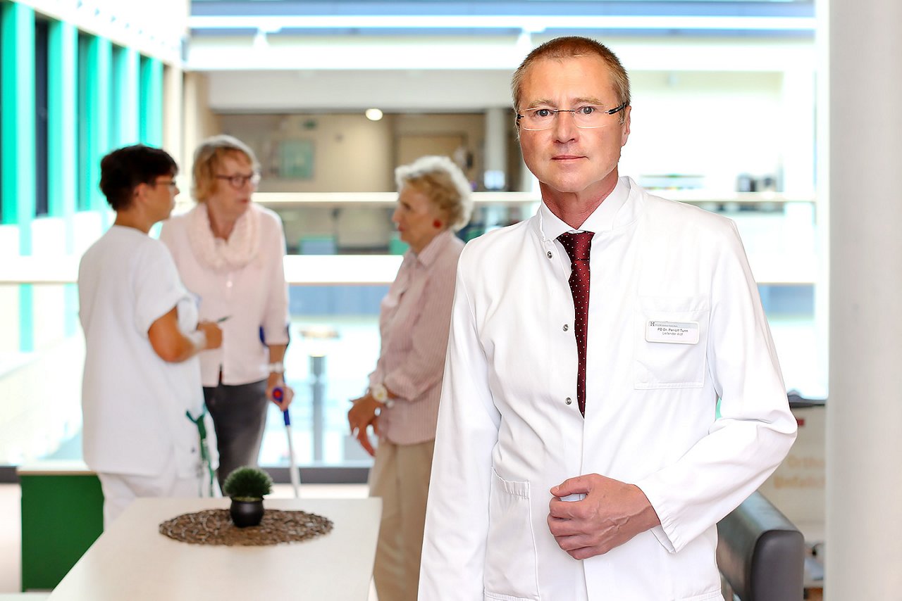 Chefarzt der Tumororthopädie auf dem Stationsflur stehend im Hintergrund zwei Patientinnen und eine Pflegekraft im Gespräch