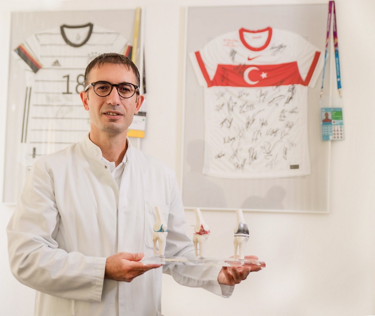 Chefarzt Dr. Ufuk Sentürk hält ein Modell mit Knieprothesen vor einer Wand mit Fußballtrikots