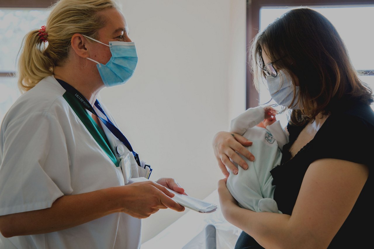 Mama mit Baby im Arm erhält von der Schwester Unterlagen überreicht.