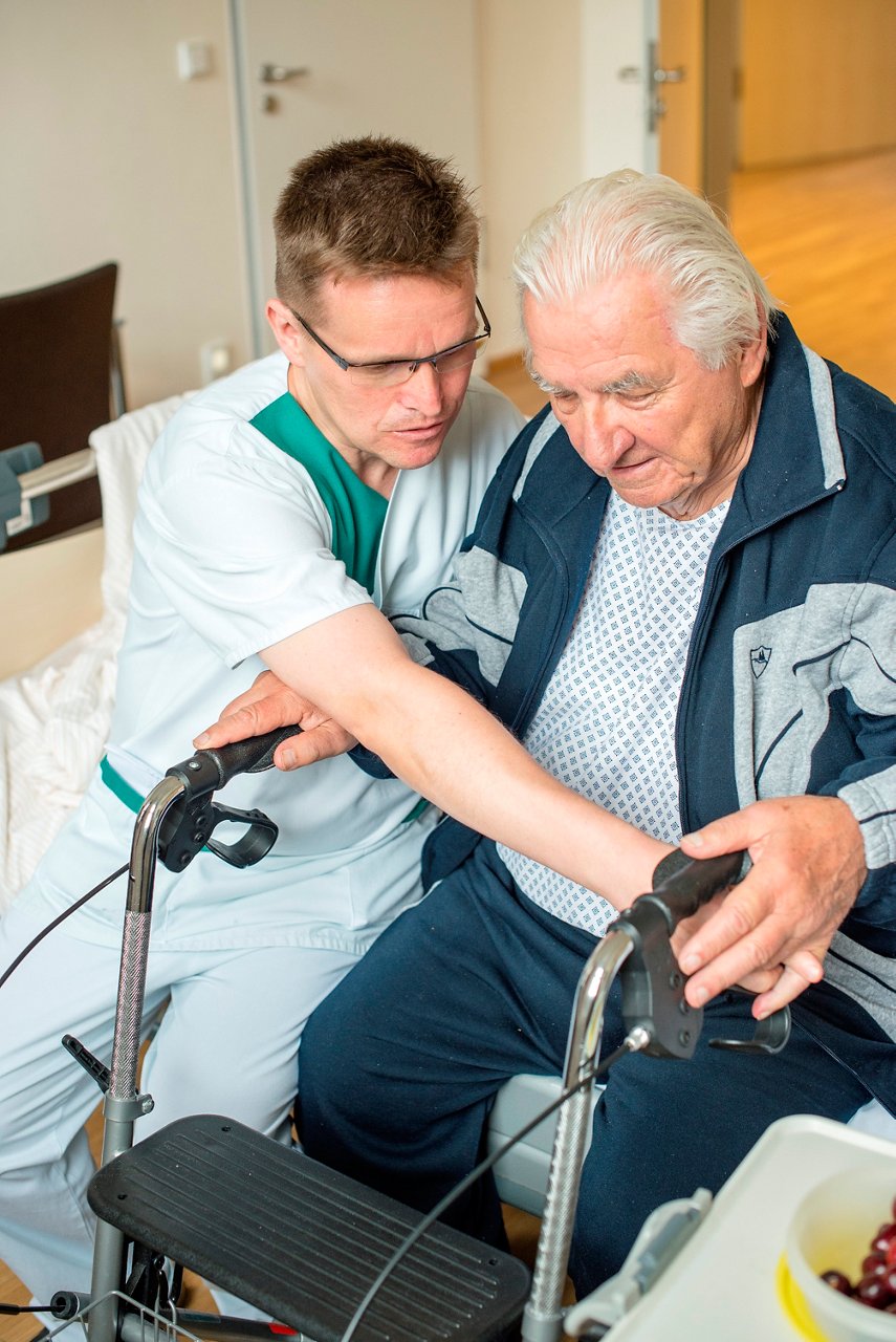 Unser freundlicher Pfleger in der Geriatrie unterstützt den Patienten beim sicheren Aufstehen mit Rollator