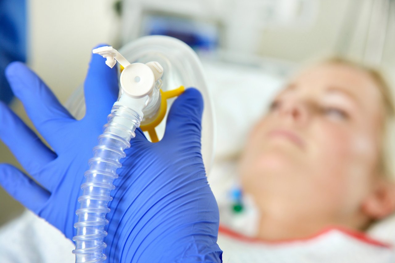 Im Vordergrund eine angeschlossene Beatmungsmaske in der Hand des medizinischen Fachpersonals im Hintergrund die Patientin im Intensivpflegebett