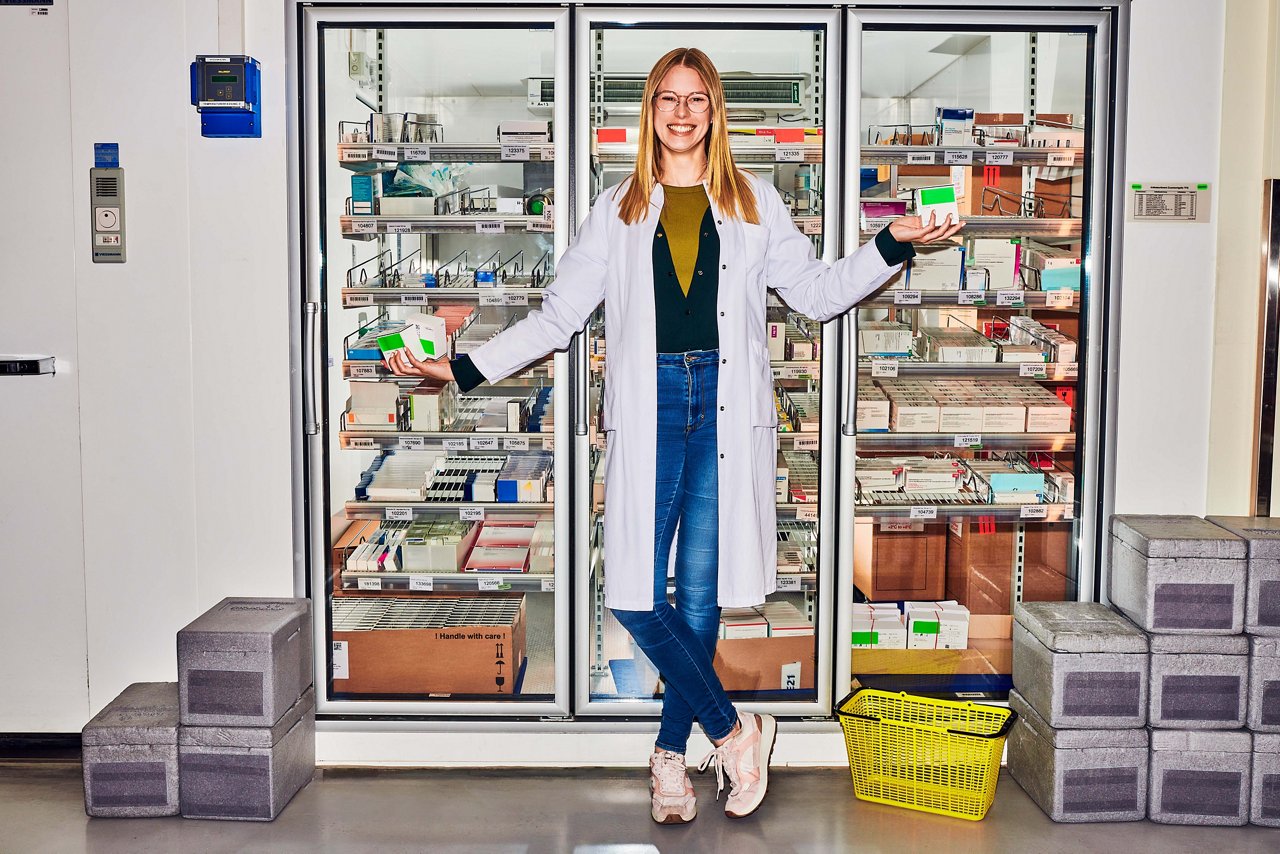 Apothekerin steht mit ausgebreiteten Armen vor einem großen Medizinschrank aus Glas und hält Medikamentenschachteln in den Händen #EchtesLeben 2023