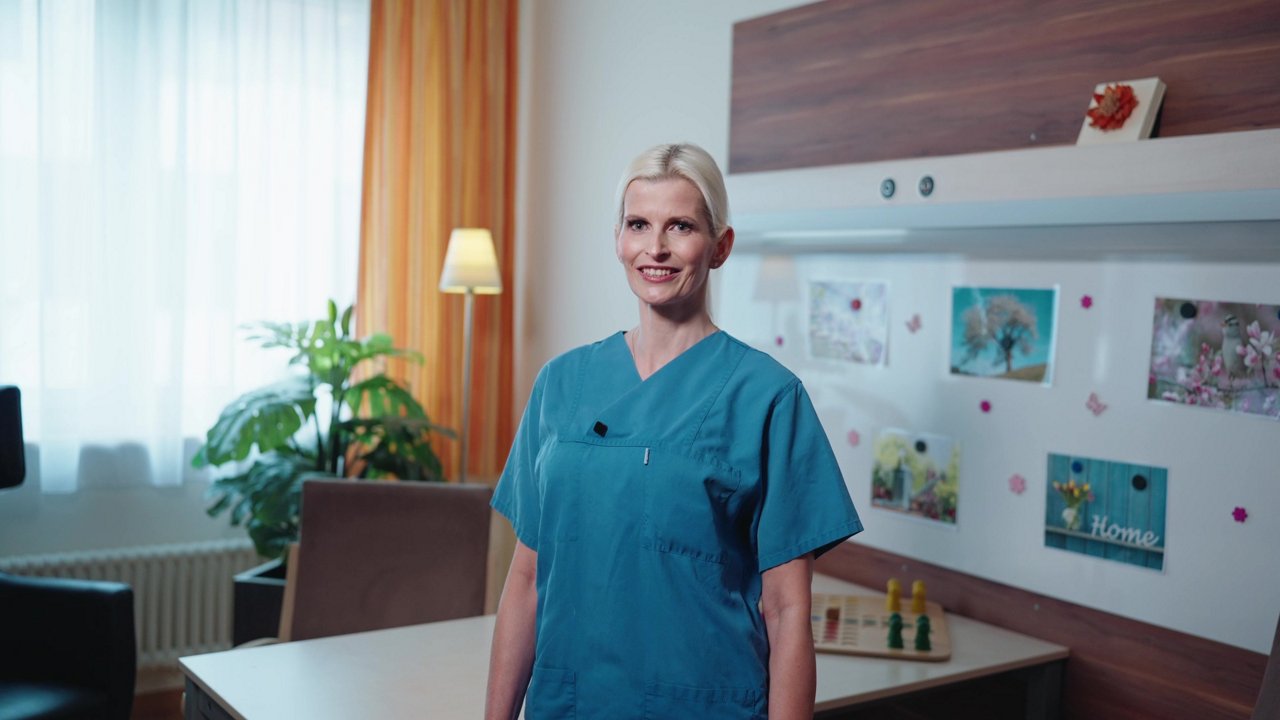 Eine freundliche Pflegekraft in blauer Uniform steht in einem hellen Raum mit Pflanzen und bunten Bildern. Der Raum strahlt Wärme und Fürsorge aus, ideal für die Pflege.