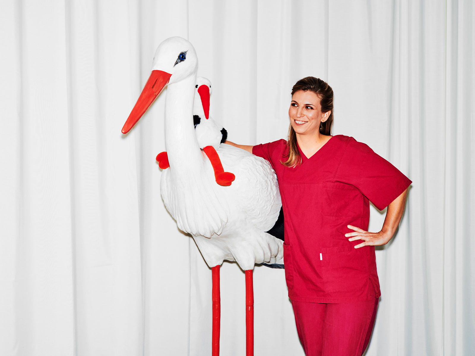 Hebamme steht mit rotem Kassack neben einer lebensgroßen Storchfigur