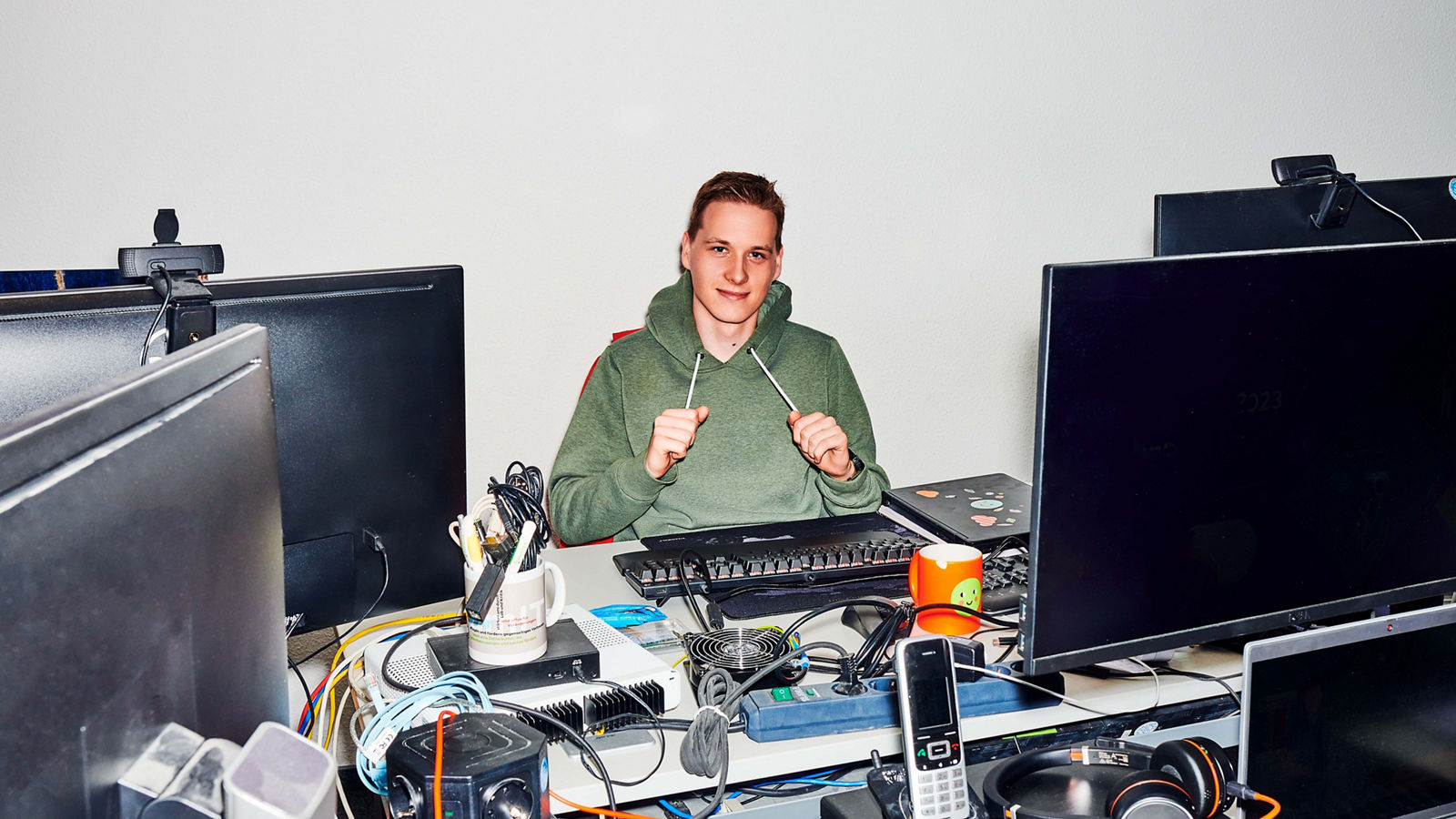 junger IT Auszubildender sitzt mit Hoody am Schreibtisch, welcher mit Monitoren, Kabeln und anderer Hardware voll gestellt ist #EchtesLeben 2023