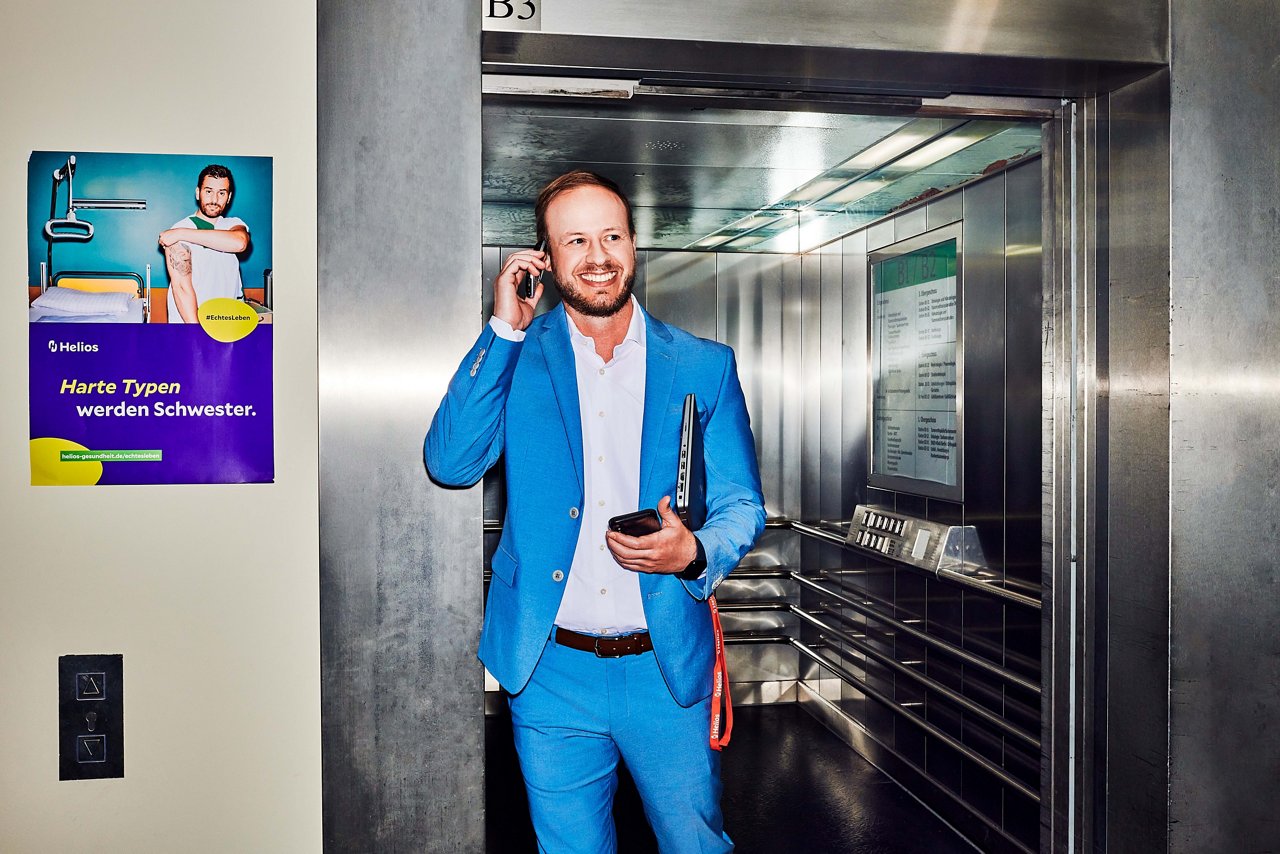 Klinikgeschäftsführer mit Telefon am Ohr und Handy und Laptop unter dem Arm vor einem geöffnetem Fahrstuhl #EchtesLeben 2023