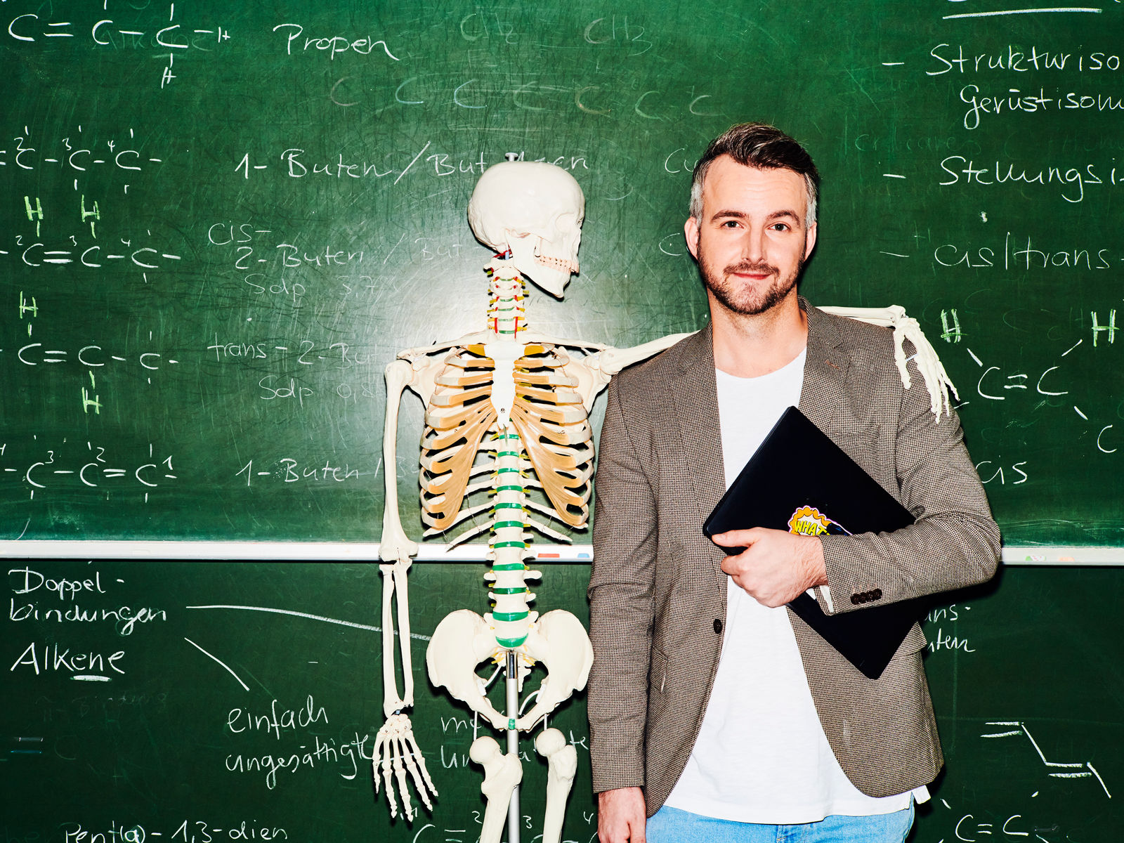 Lehrer steht vor einer Tafel ein Skelett legt seinen Arm um seine Schulter #EchtesLeben 2021/22