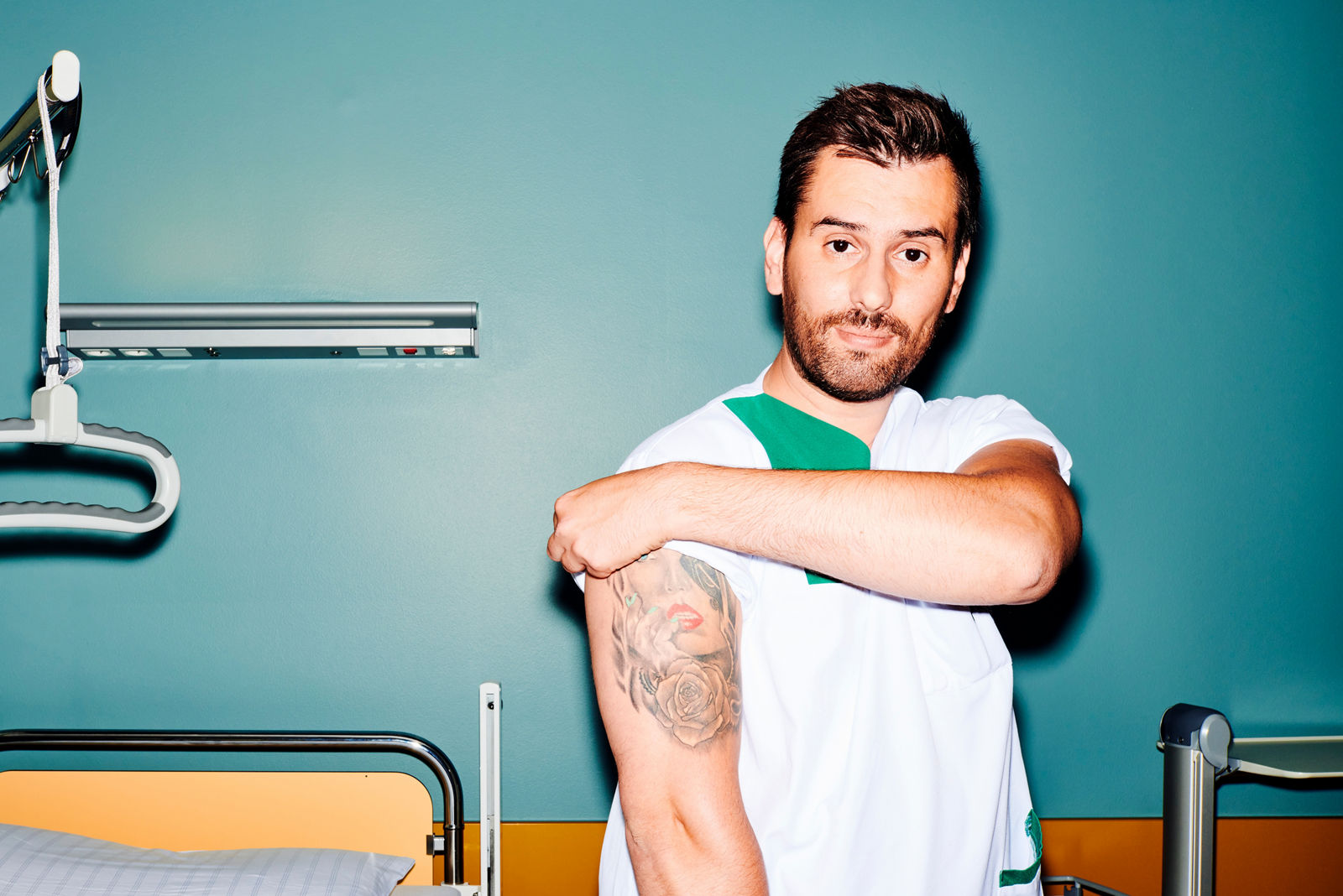 Pfleger steht am Patientenbett und krempelt sich einen Ärmel hoch, dabei wird sein Tattoo am Oberarm sichtbar #EchtesLeben 2021/22