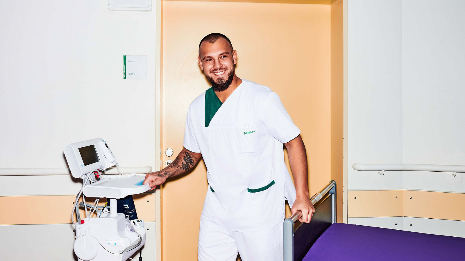 lächelnder Krankenpflegehelfer auf Stationsflur mit Patientenbett und Patientenmonitor #EchtesLeben 2023