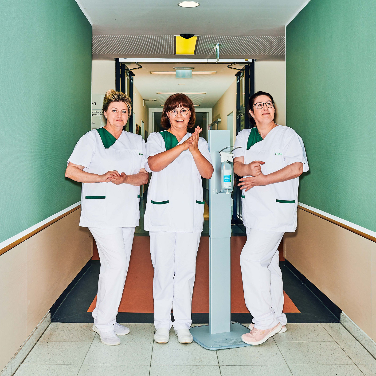3 Hygiene Mitarbeiterinnen desinfizieren sich die Hände am Desinfektionsspender im Klinikflur #EchtesLeben 2023