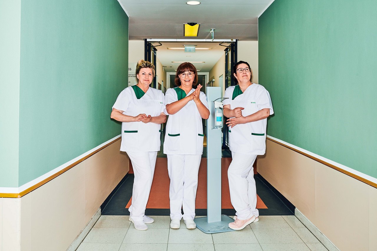 3 Hygiene Mitarbeiterinnen desinfizieren sich die Hände am Desinfektionsspender im Klinikflur #EchtesLeben 2023