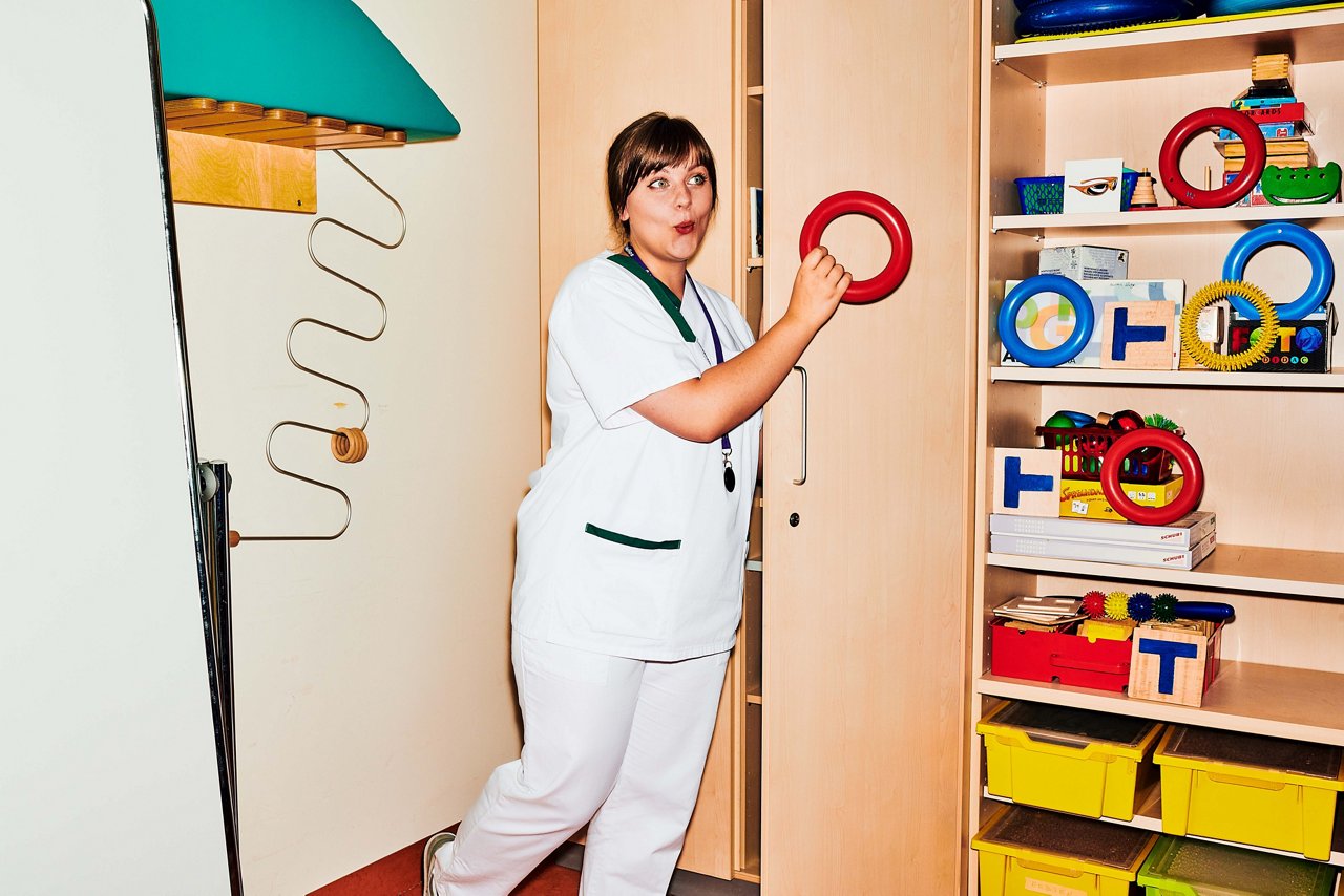 Logopädin steht vor Schrank mit vielen, bunten Hilfsmitteln im Therapieraum und formt mit dem Mund ein O während sie einen Gummiring hält #EchtesLeben