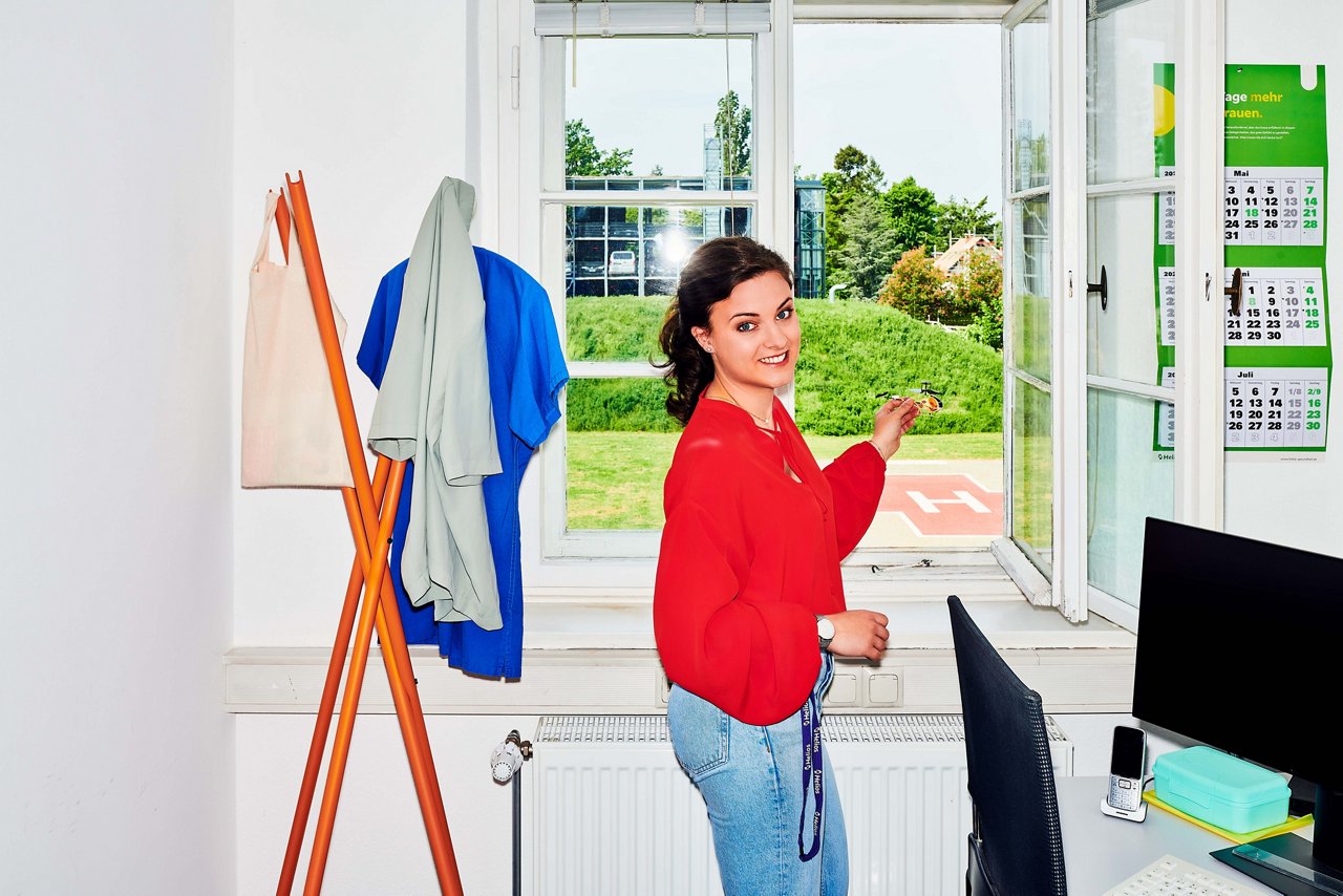 Trainee steht vor einem Fenster - im Hintergrund der Hubschrauberlandeplatz der Klinik #EchtesLeben 2023