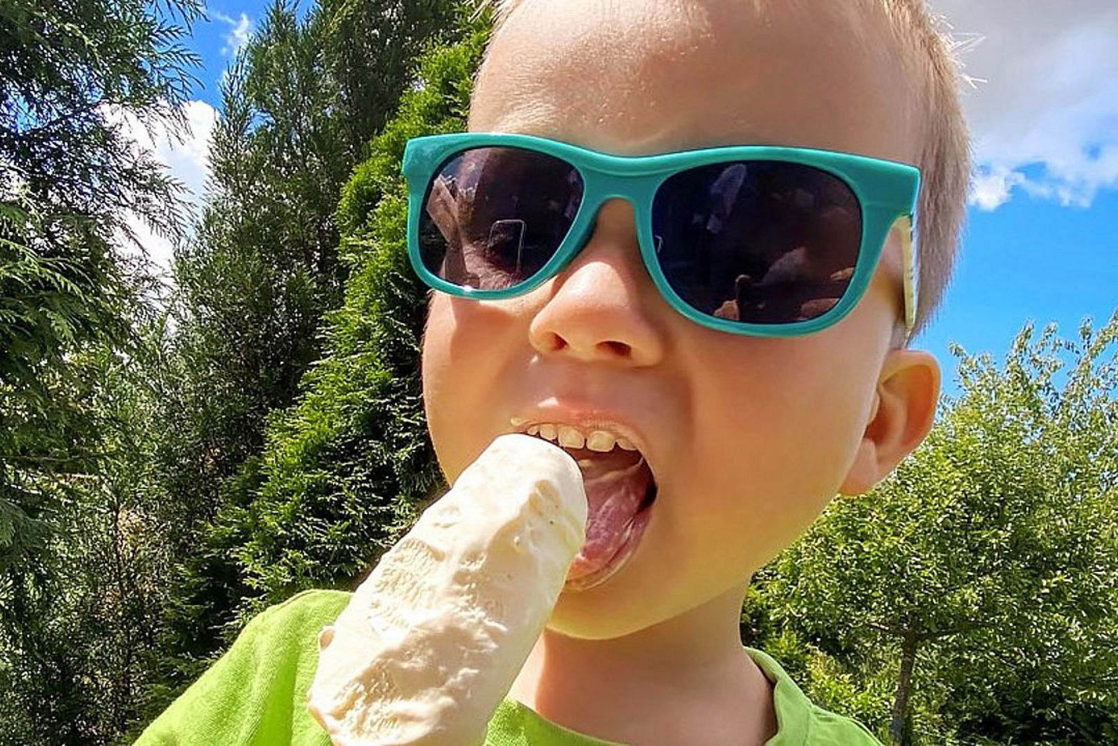 Ein kleiner Junge im Vorschulalter in einem grünen T-shirt mit petrolfarbiger Sonnenbrille leckt an einem Sommertag ein Eis am Stiel.