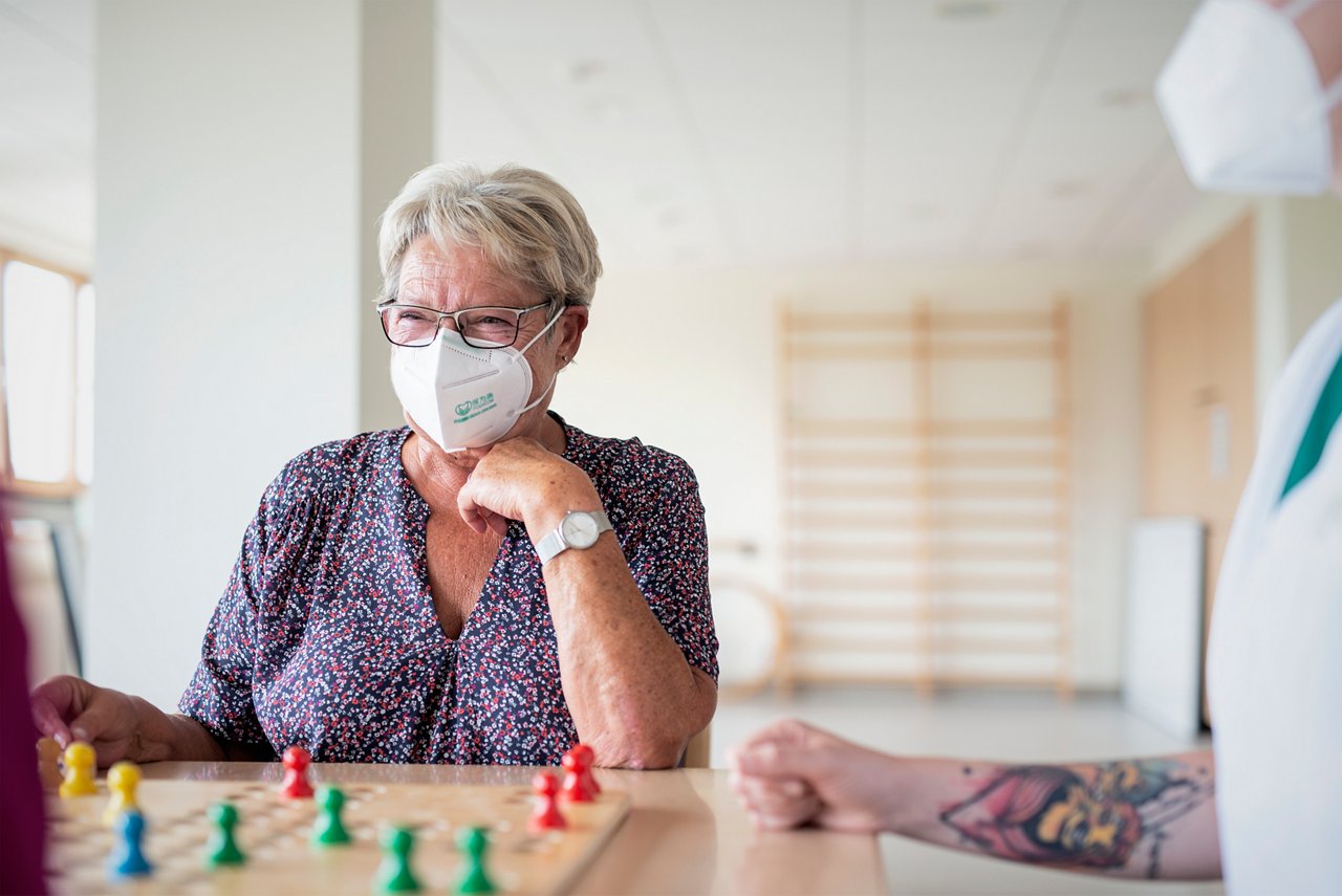 Geriatrische Patientin mit FFP2-Maske spielt Brettspiel 