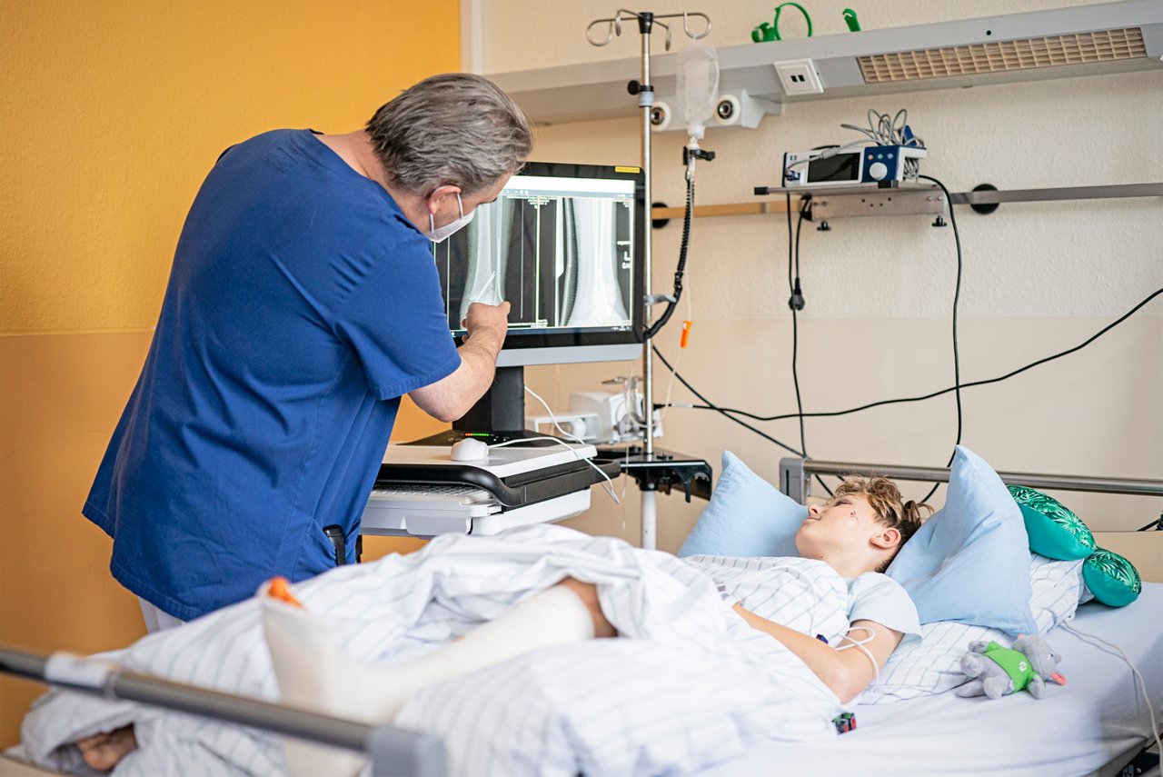 Chefarzt Dr. Uhlig  erklärt einem Jungen, der mit Gipsbein im Patientenbett liegt,  die Röntgenbilder