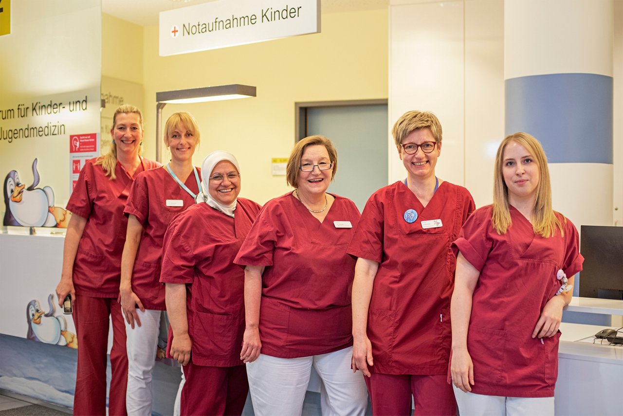 Pflegeteam der Kinderklinik Notfall Ambulanz vorm Stationstresen in rotem Kasack