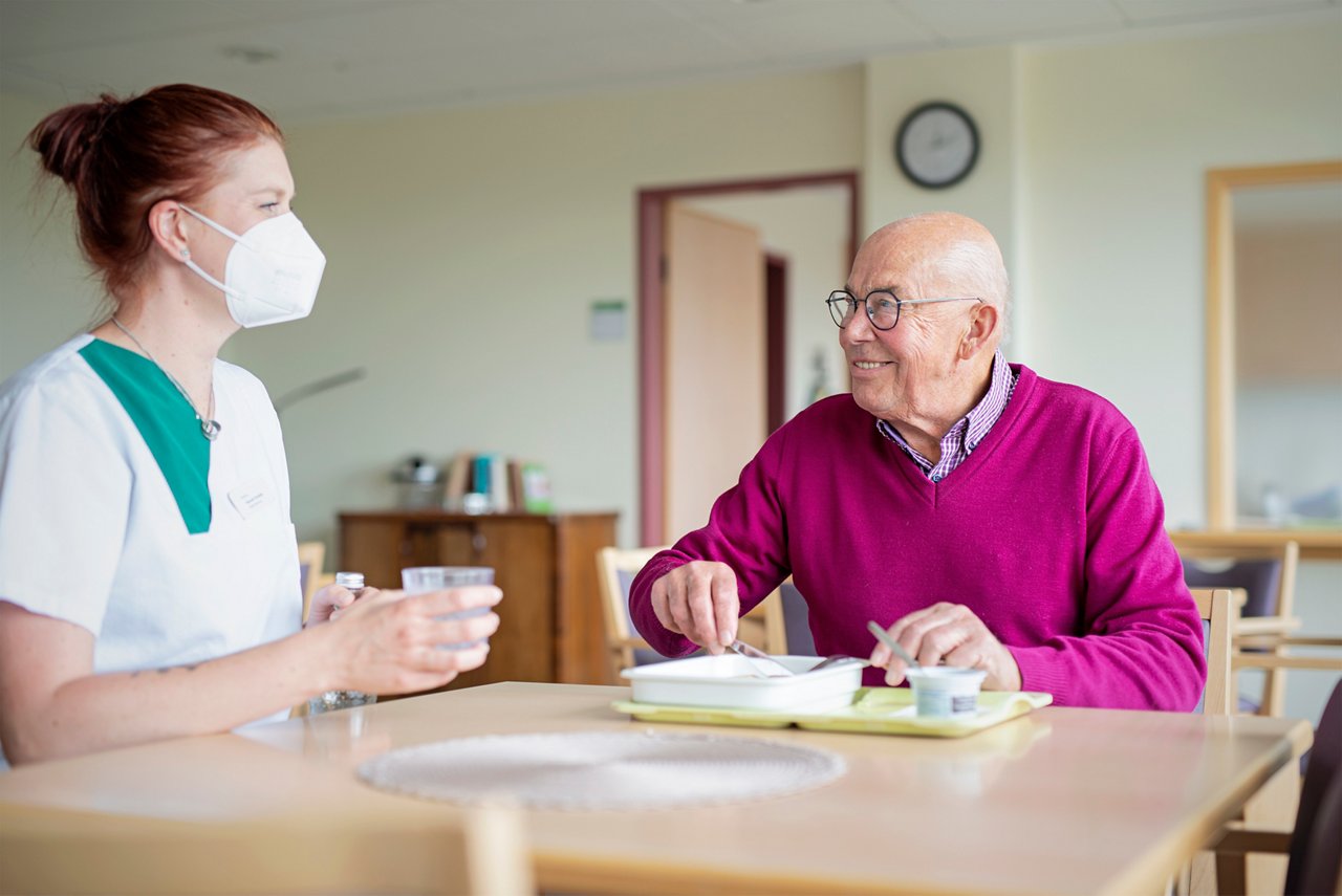 Lächelnder geriatrischer Patient mit Brille beim Essen mit Pflegerin