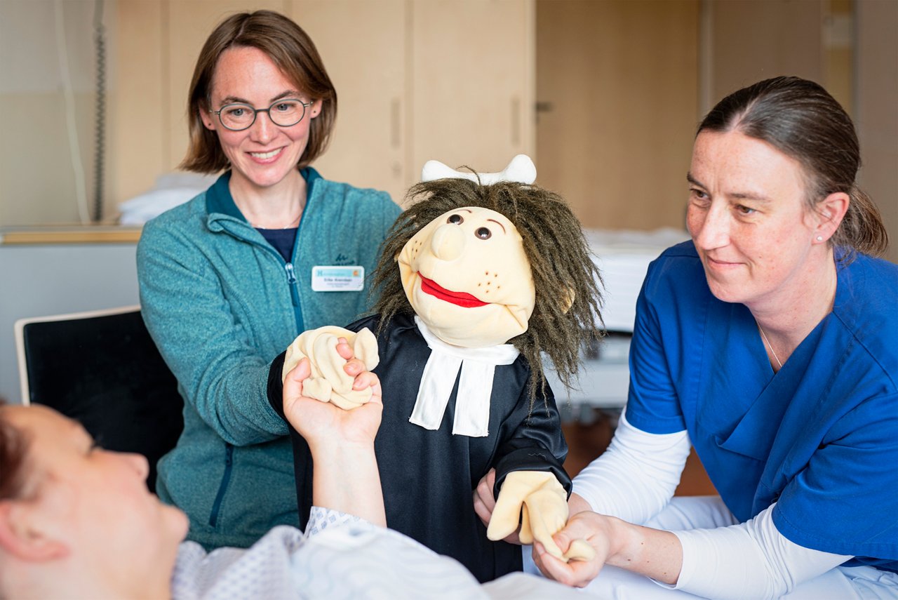 Eine Ärztin und eine Seelsorgerin sprechen mit einer Patientin auf der Palliativstation - unterstützt von einer Therapiepuppe..