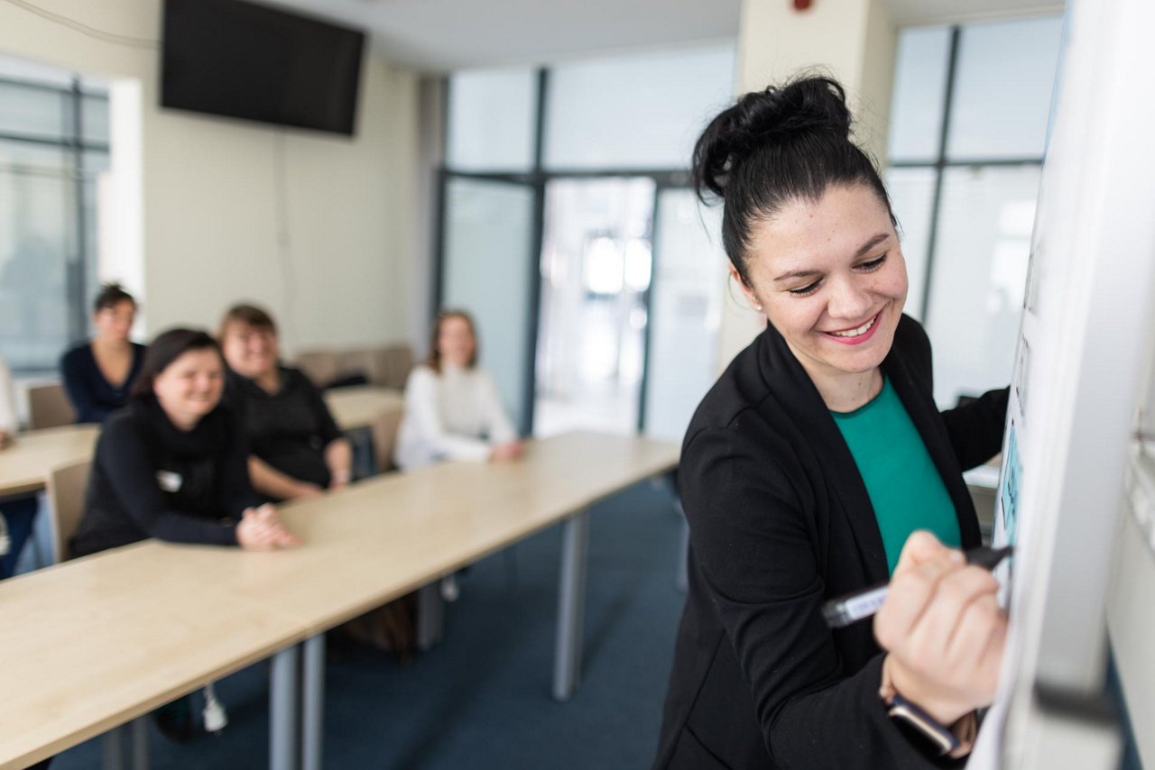 Eine engagierte Trainerin präsentiert in einem Seminarraum, während die Teilnehmerinnen aufmerksam zuhören. Der Fokus liegt auf interaktiven Lernmethoden und Teamarbeit.